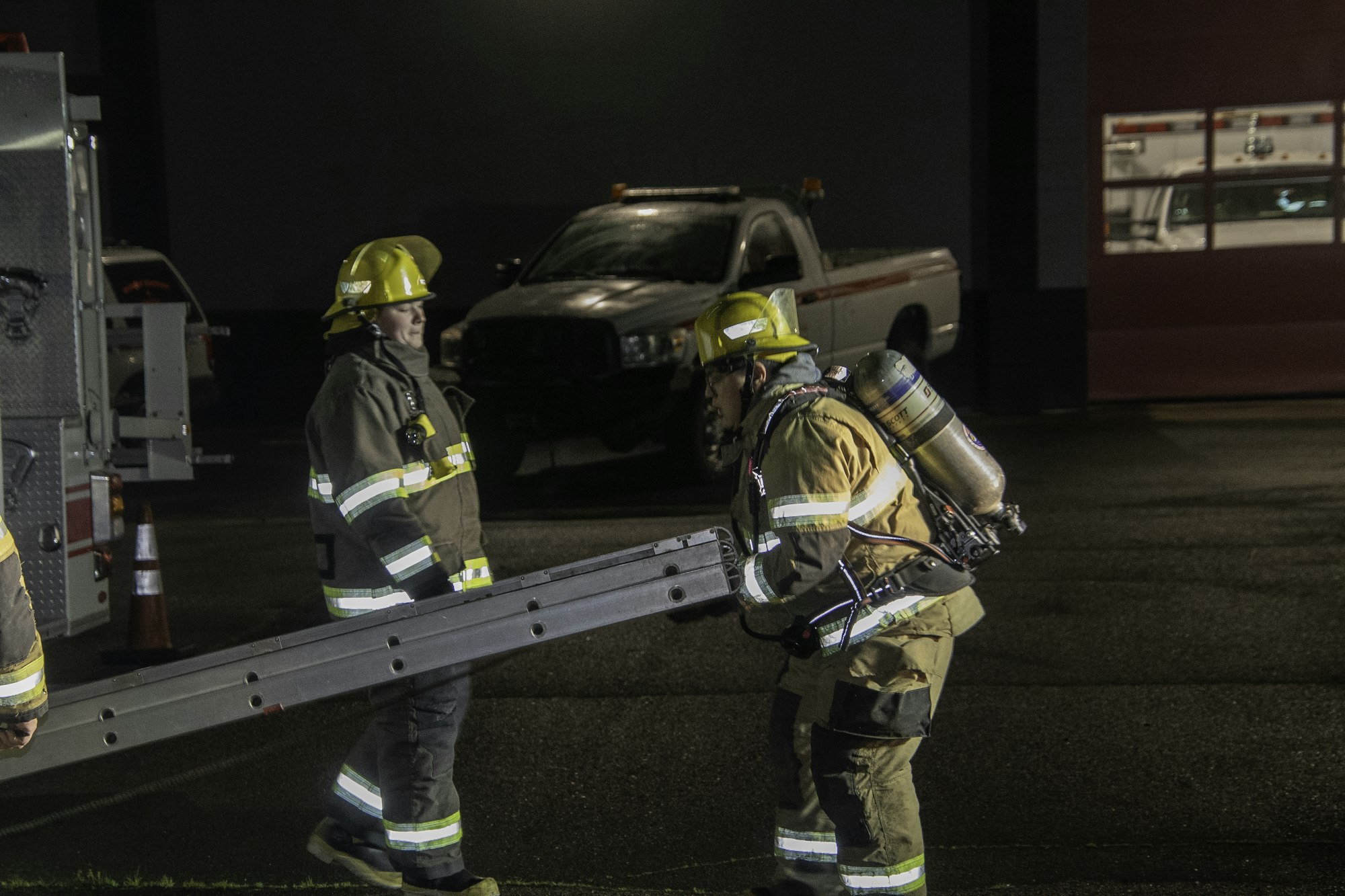 Two firefighters are working together at night, one carrying a ladder while wearing gear, with a fire truck and vehicle in the background.