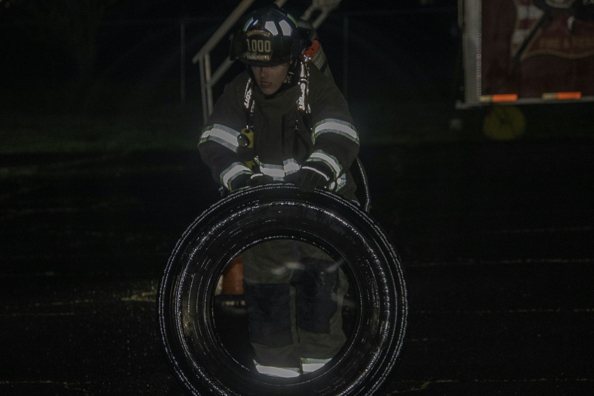 A firefighter in gear is carrying a large tire at night, focusing on their task under dim lighting.