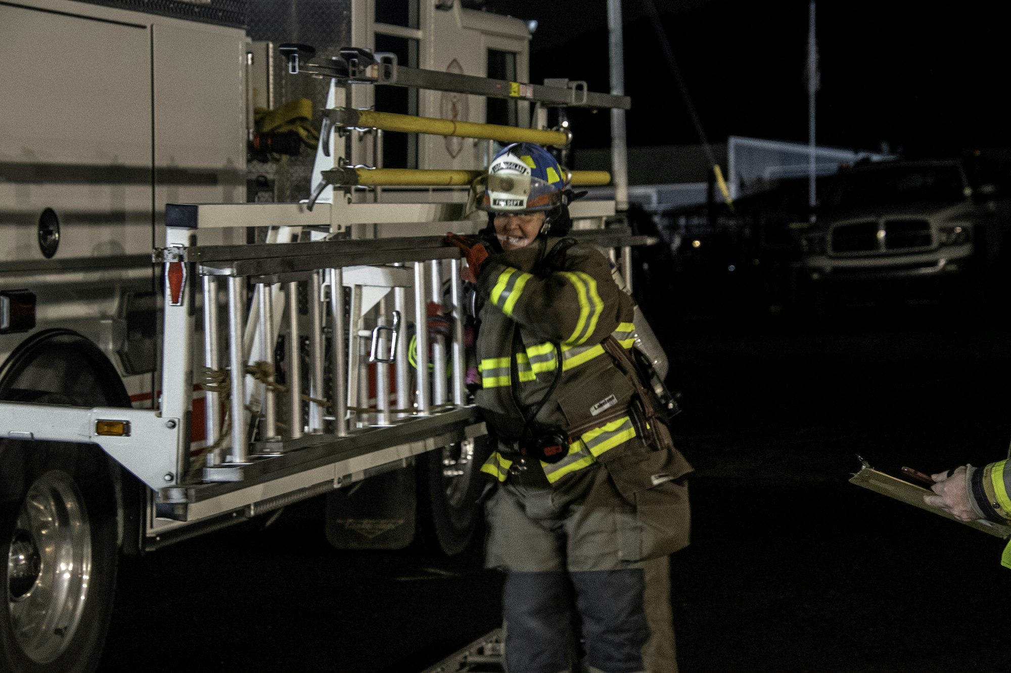 A firefighter in gear is handling a ladder at night, while another individual appears to be holding a clipboard nearby.