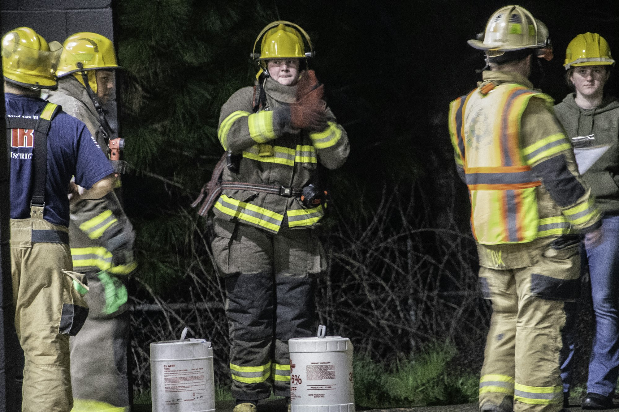 A group of firefighters in uniforms and helmets are gathered, with one clapping and others engaged in conversation.