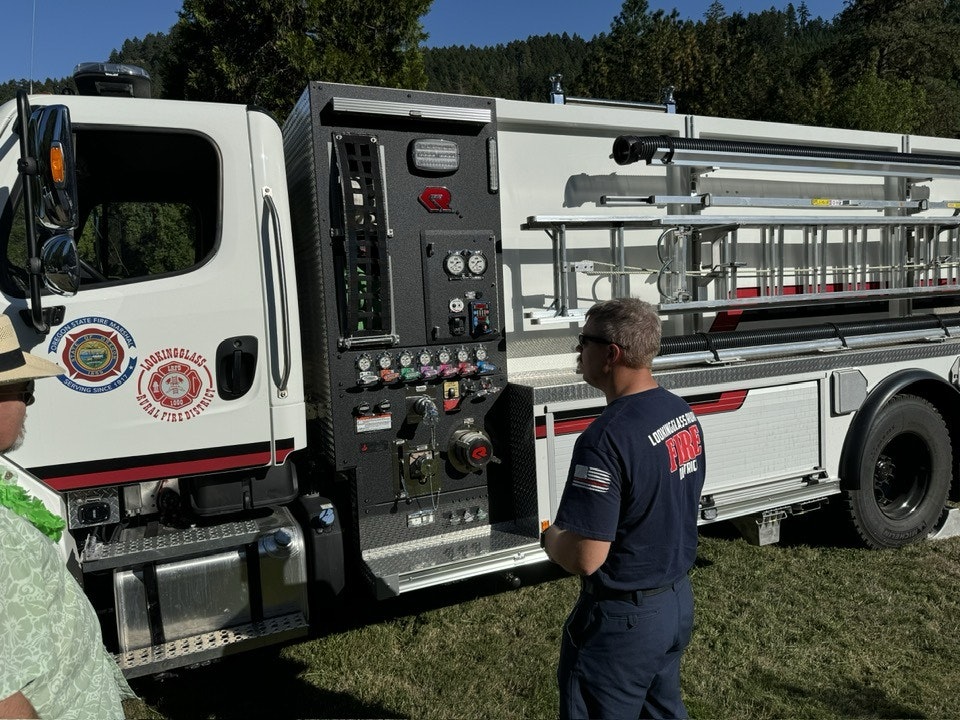 A firefighter near a fire truck control panel, with hoses and equipment visible.