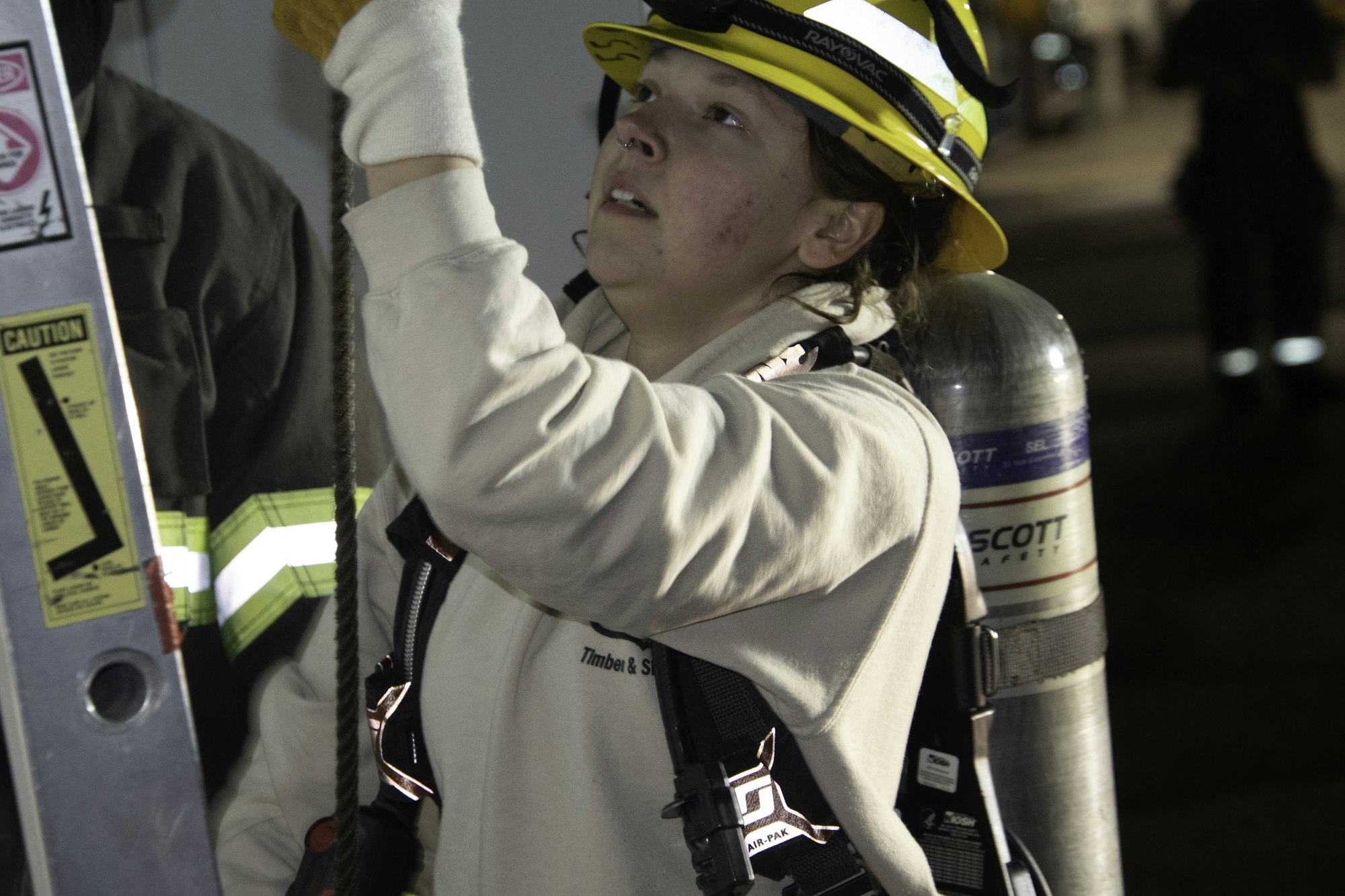 A firefighter in gear operates a rope, focused on a task, with a ladder and other personnel in a dimly lit setting.