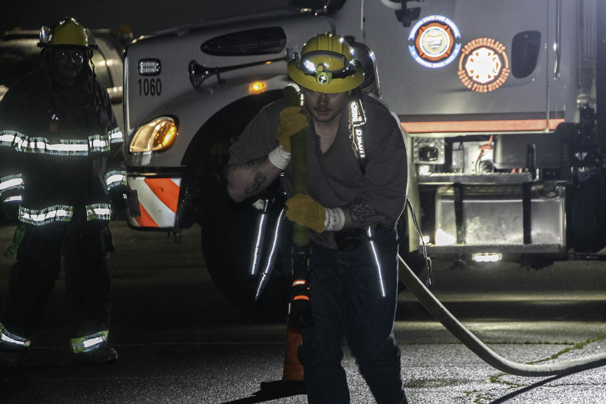 A firefighter in gear carries a hose towards a fire truck at night, while another firefighter stands nearby.