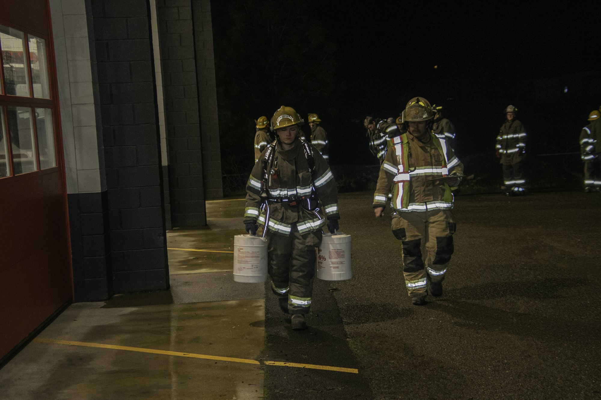 Firefighters in gear are carrying buckets outside a fire station at night, possibly preparing for a training exercise.