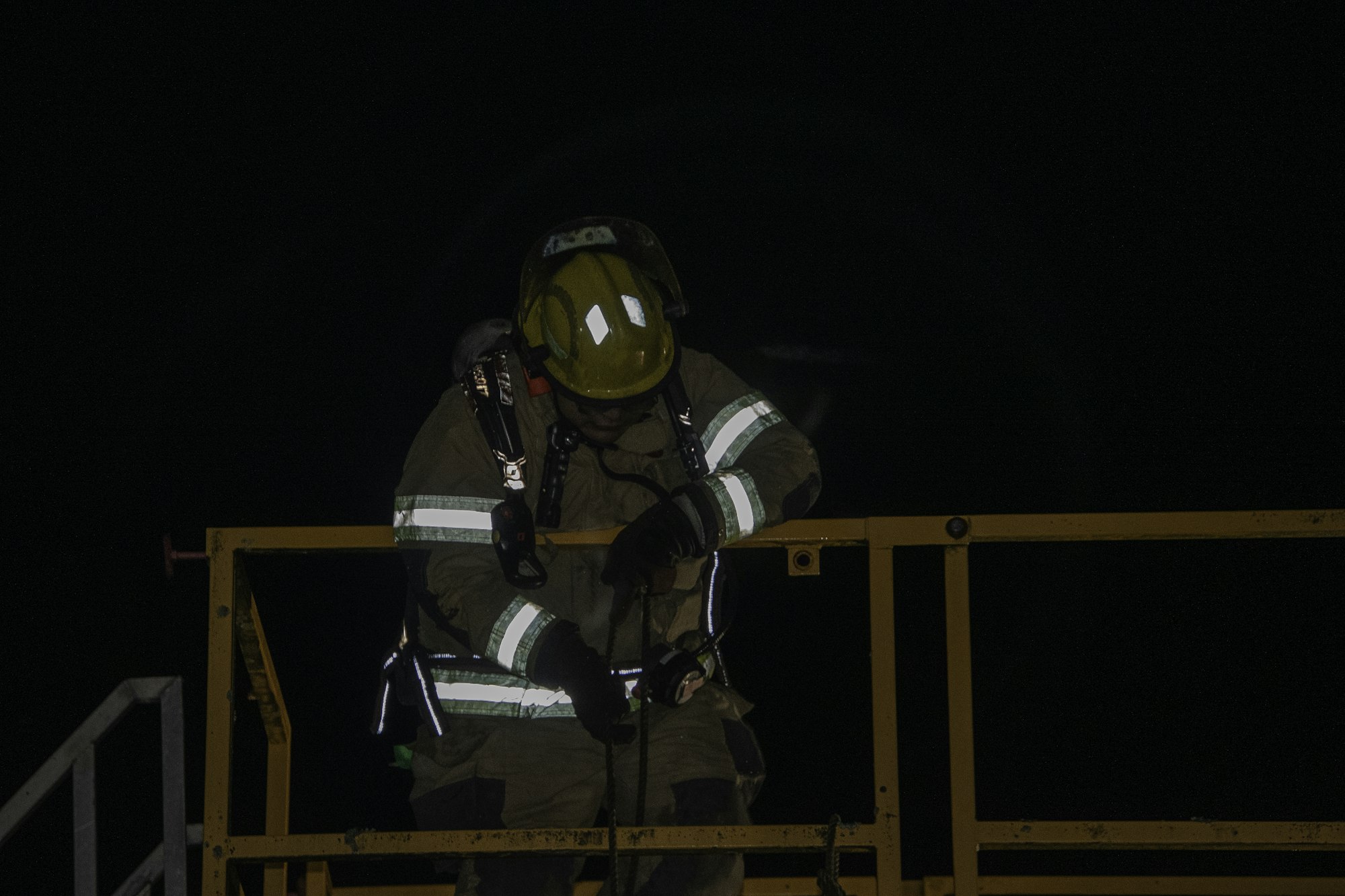 A firefighter, dressed in gear and a helmet, is working on a platform in a dark environment, focused on a task.