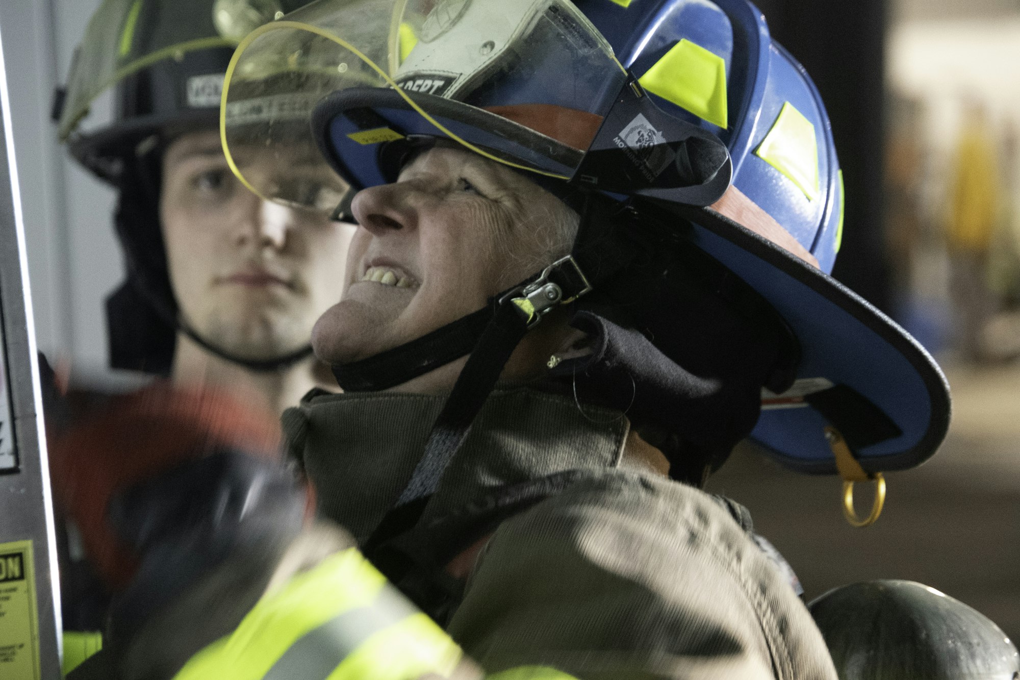 The image shows two firefighters, one focused on equipment while the other looks on. Both are in protective gear.