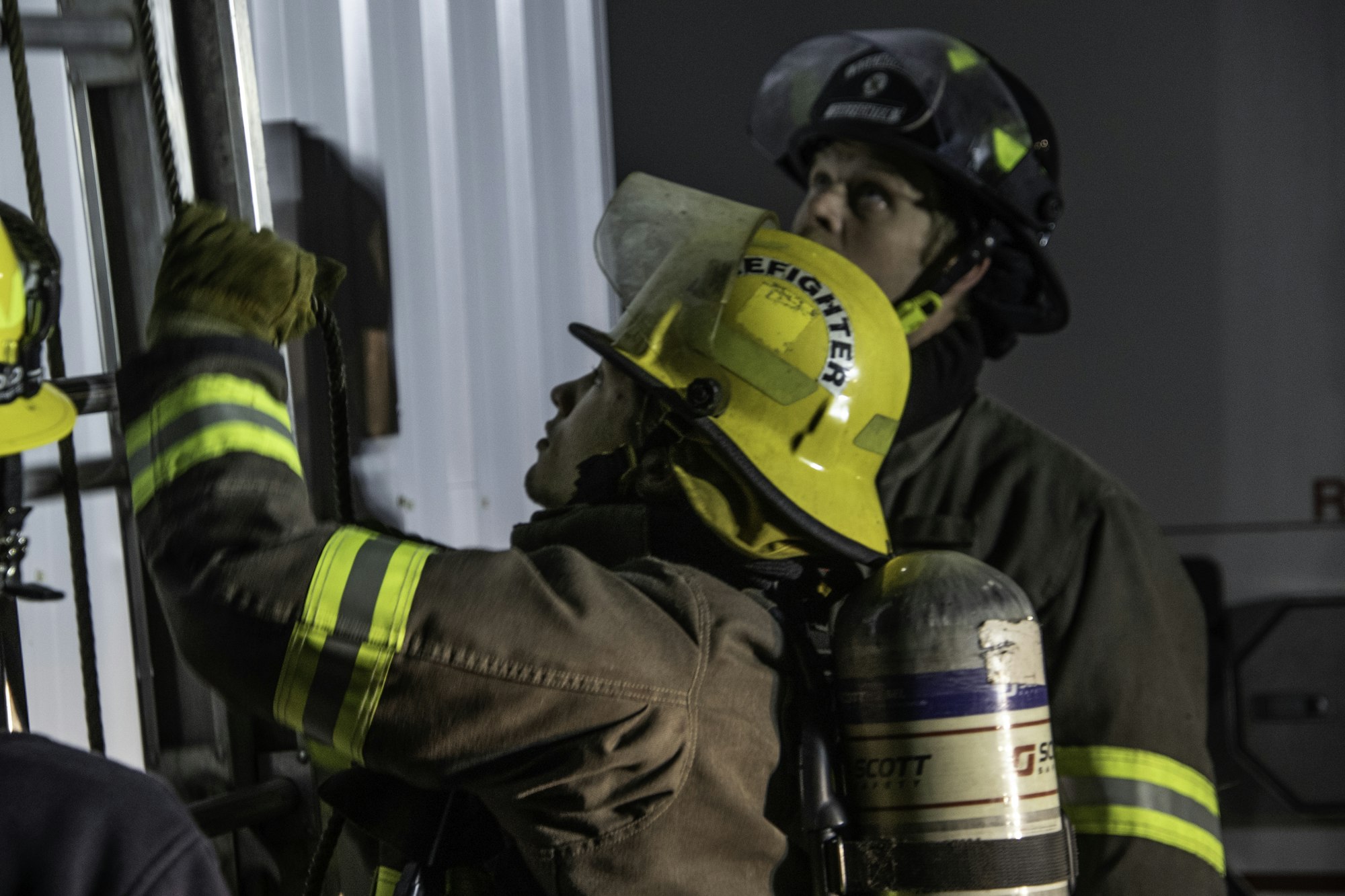 Two firefighters in gear are looking up, one adjusting a rope on a ladder, emphasizing teamwork and preparation for a task.