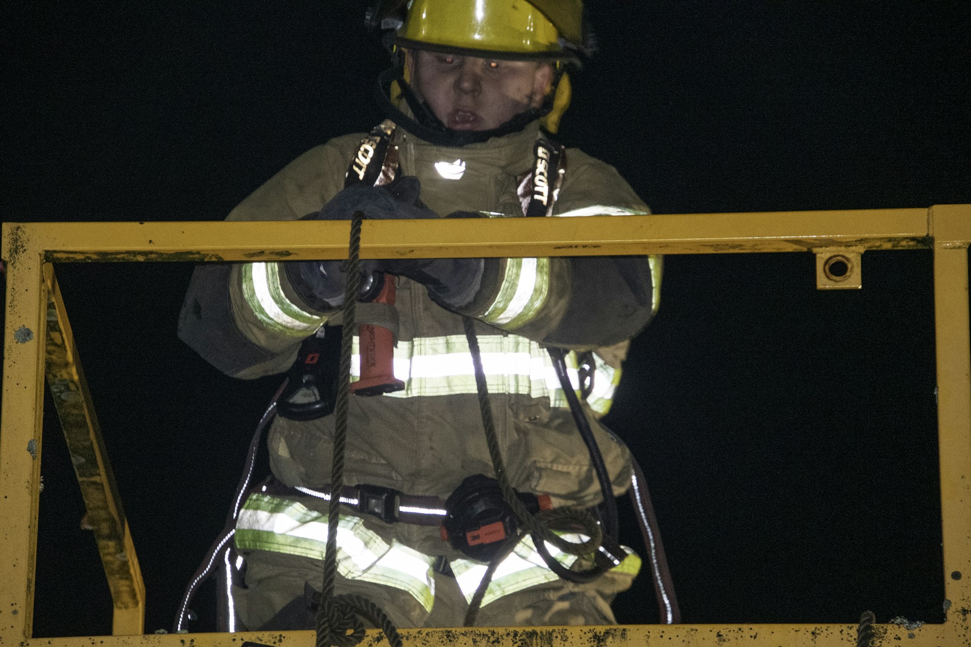 A firefighter in gear works at night, focusing on a rope while on a raised platform, highlighting safety and rescue efforts.