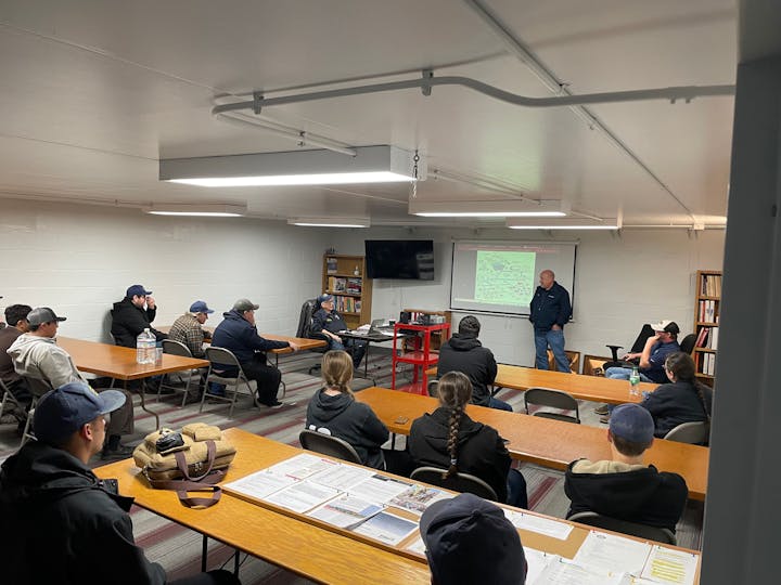 A group meeting in a classroom-like setting, with a speaker presenting in front of an audience seated at tables.