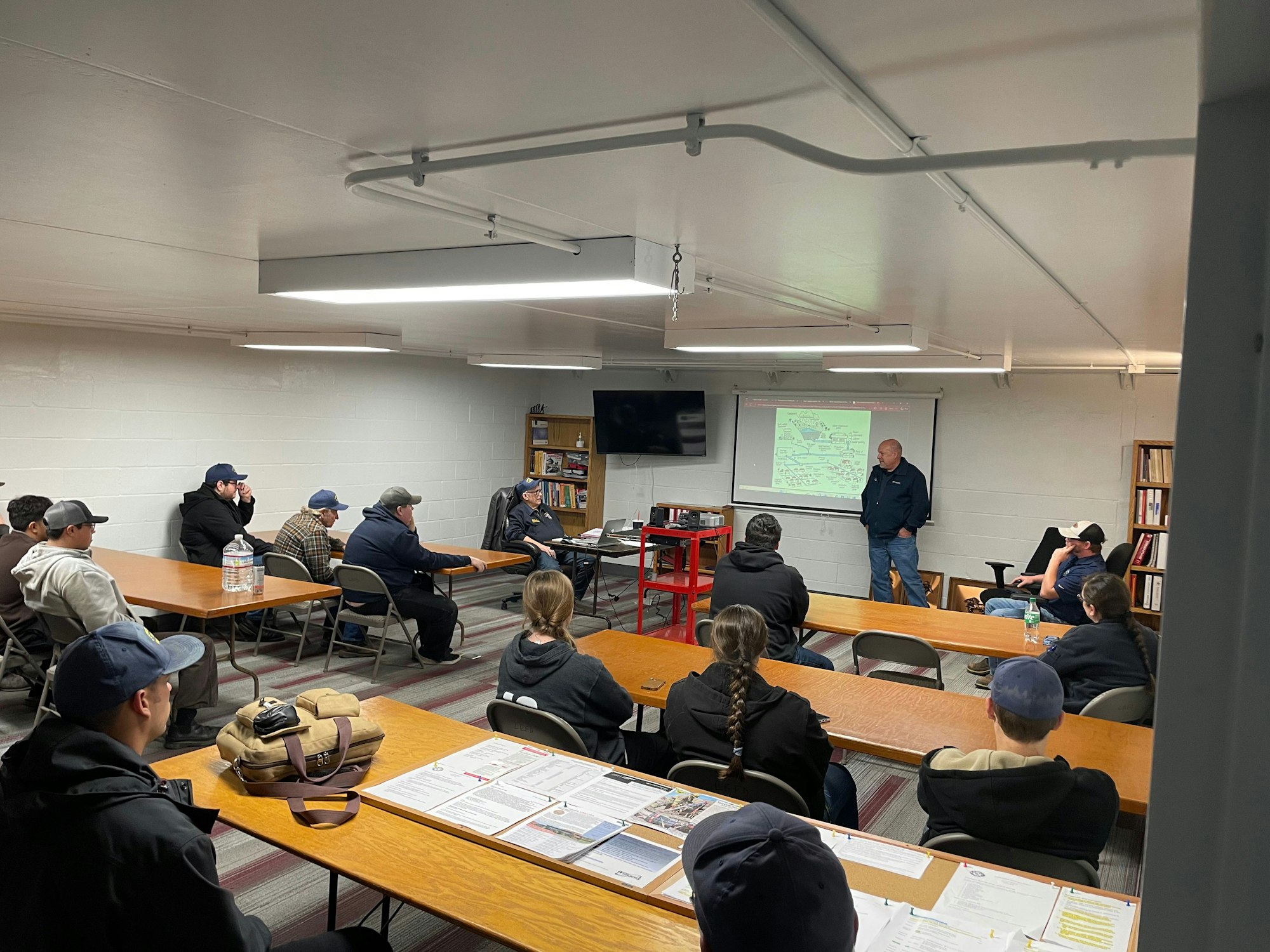 A group meeting in a classroom-like setting, with a speaker presenting in front of an audience seated at tables.
