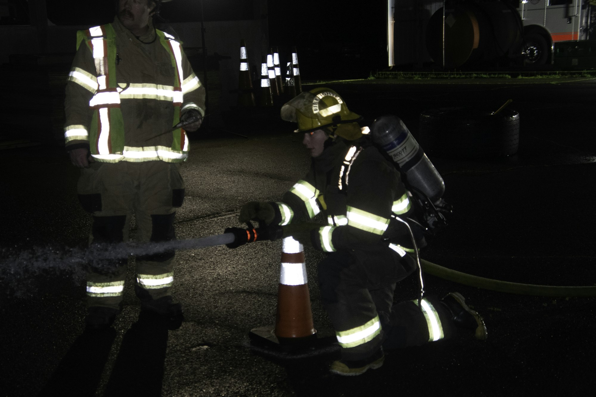 Two firefighters in protective gear are practicing with a fire hose at night, one is kneeling and spraying water.