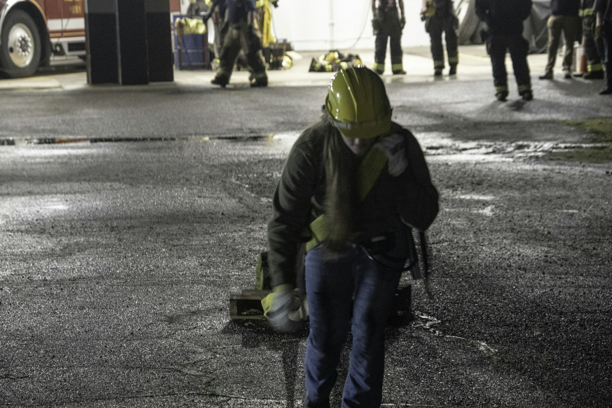 A person in a hard hat walks on a wet surface, with emergency responders and equipment visible in the background.