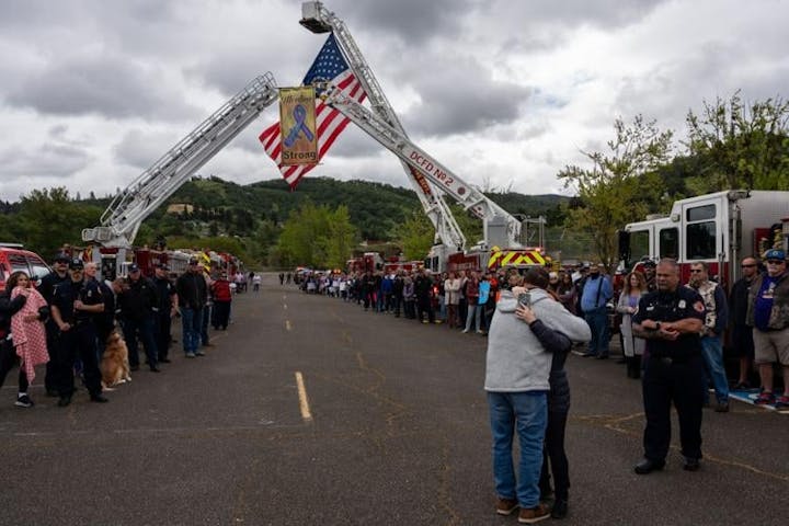 A crowd stands by fire trucks with raised ladders displaying a flag. Two people hug in the foreground.