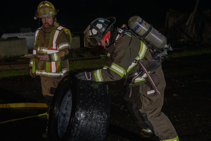 Two firefighters in gear, one holding a clipboard and the other inspecting a tire, possibly during a training exercise at night.