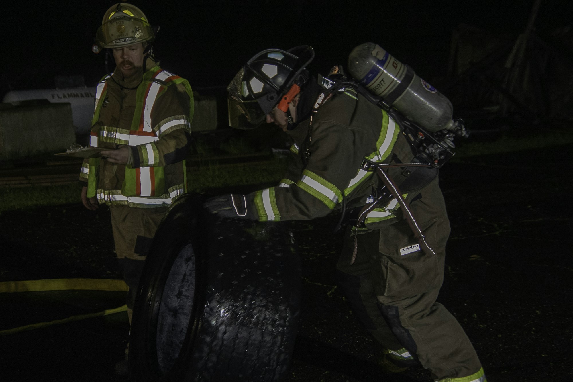 Two firefighters in gear, one holding a clipboard and the other inspecting a tire, possibly during a training exercise at night.