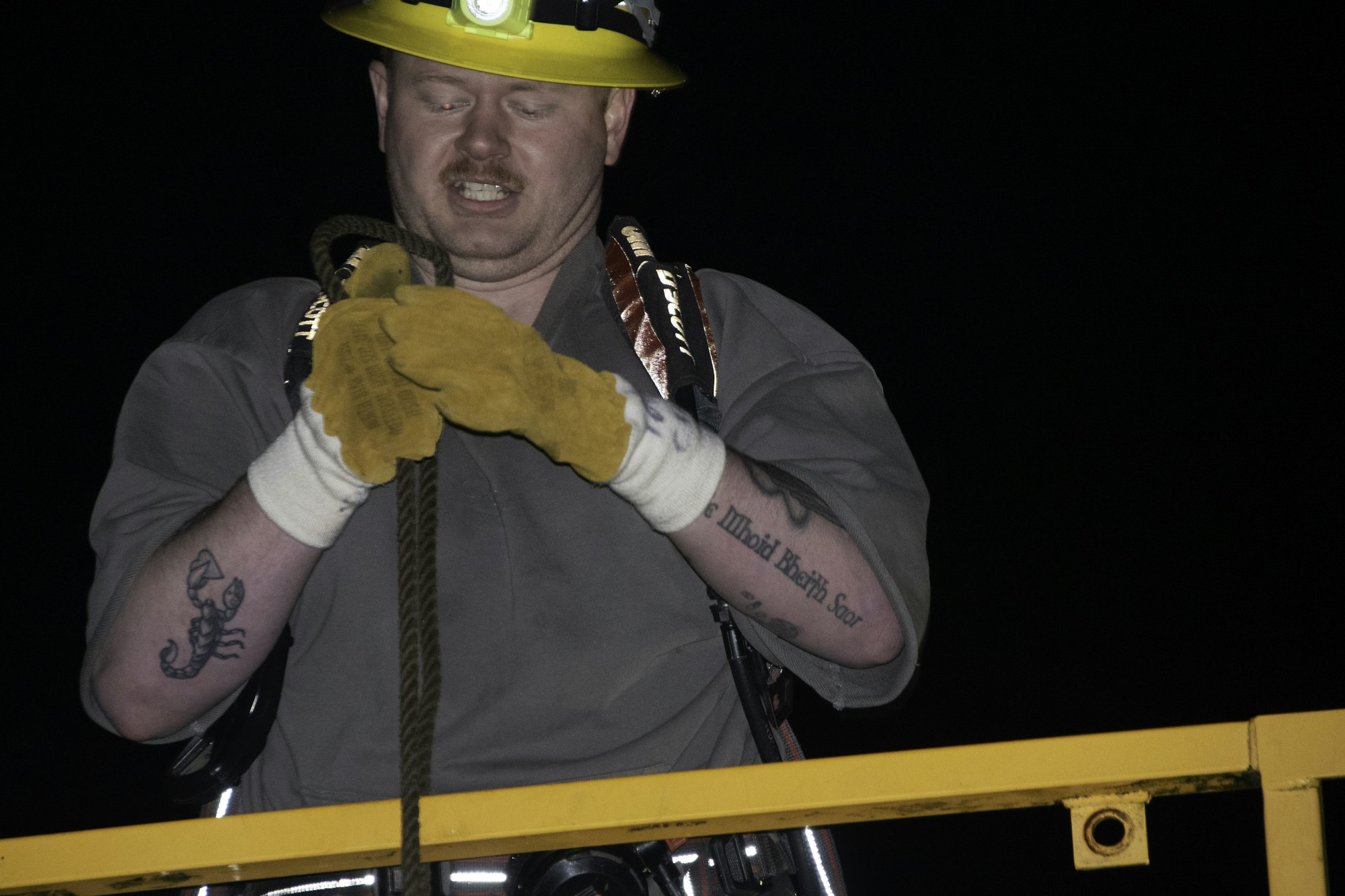 A person with a headlamp and gloves is handling a rope, wearing a work shirt, with a visible tattoo on their arm.