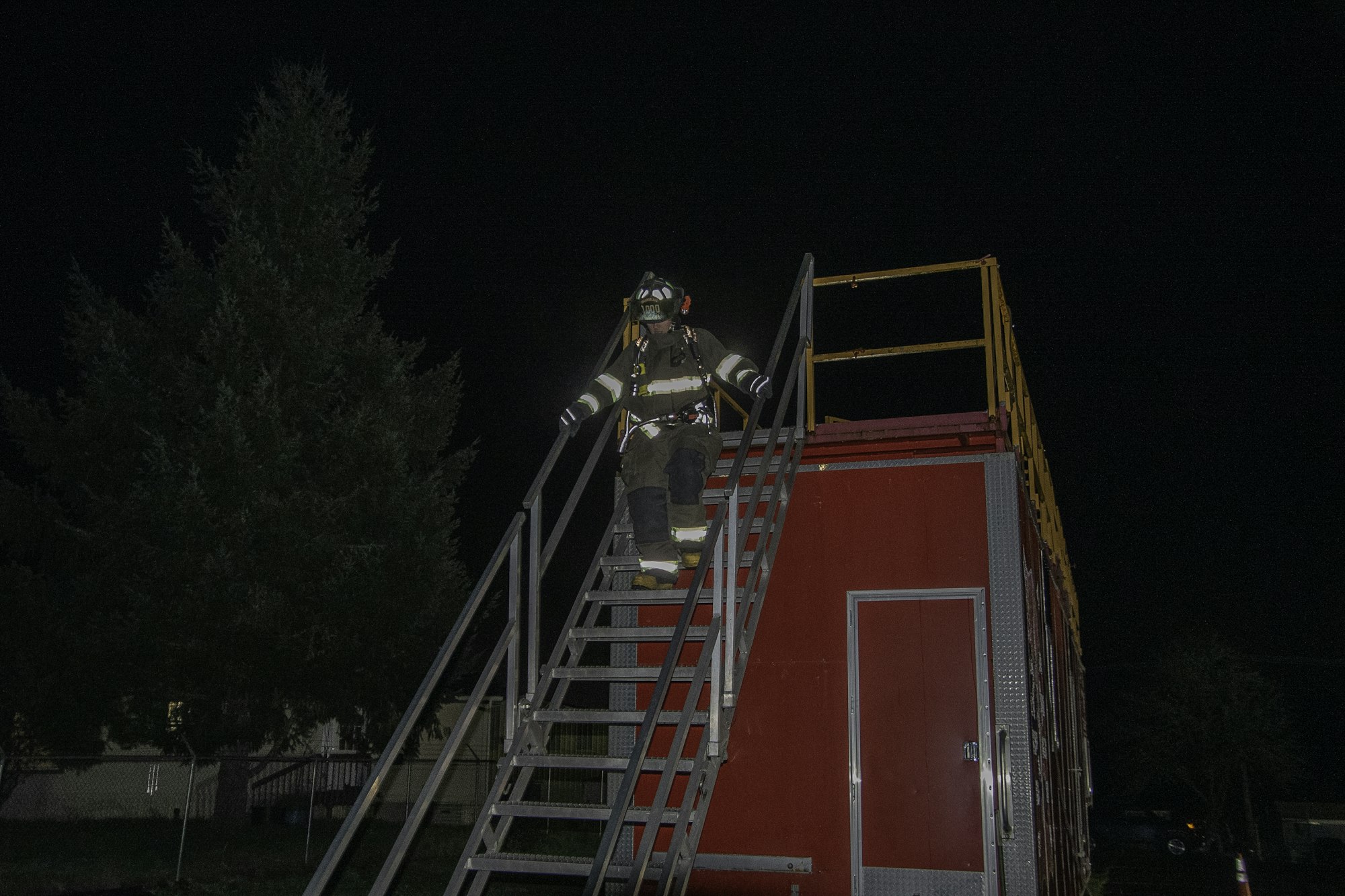 A firefighter descends a metal staircase beside a red training structure at night, surrounded by trees.