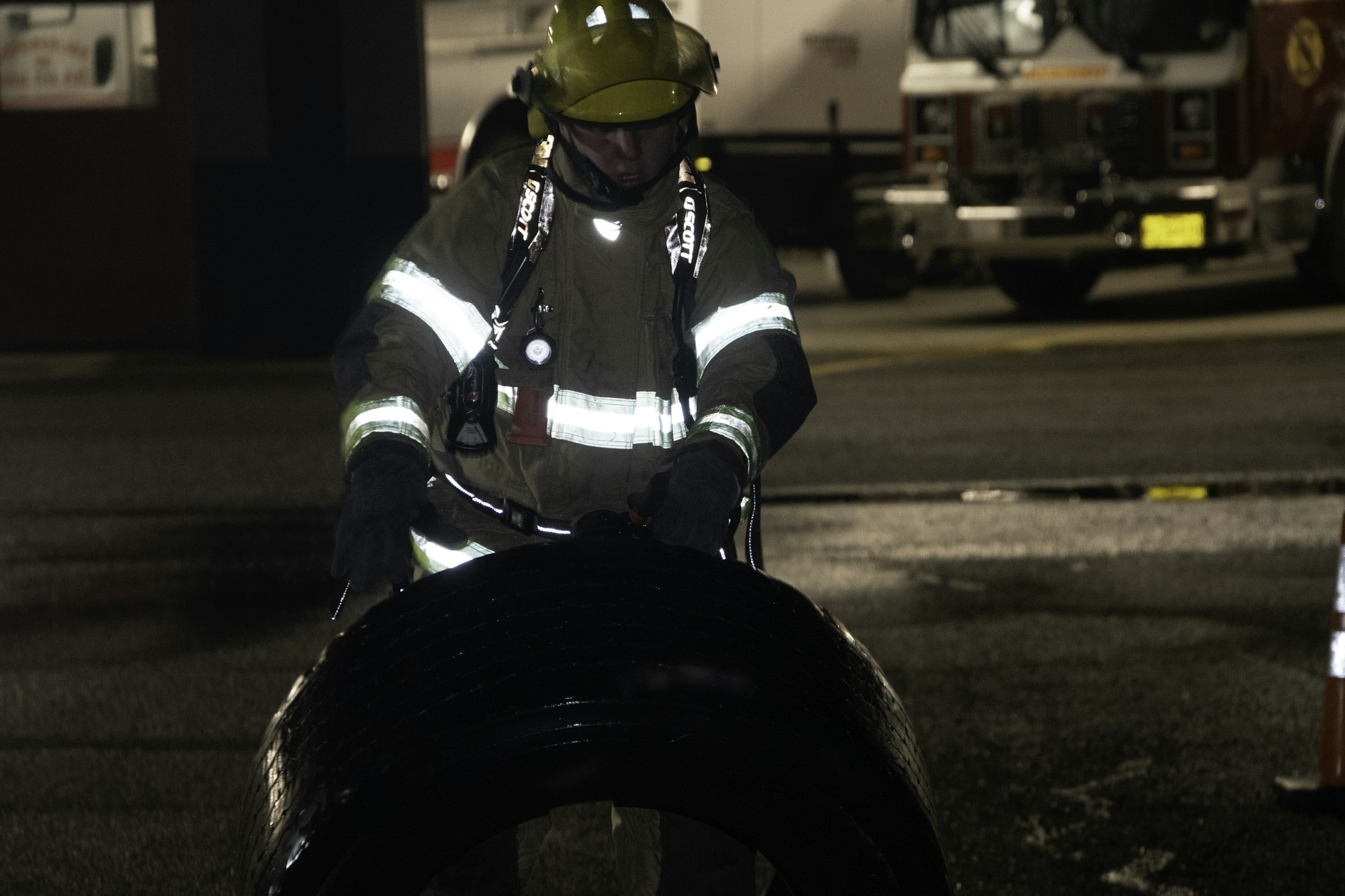 A firefighter in gear handling a large tire, with emergency vehicles in the background, during a nighttime operation.