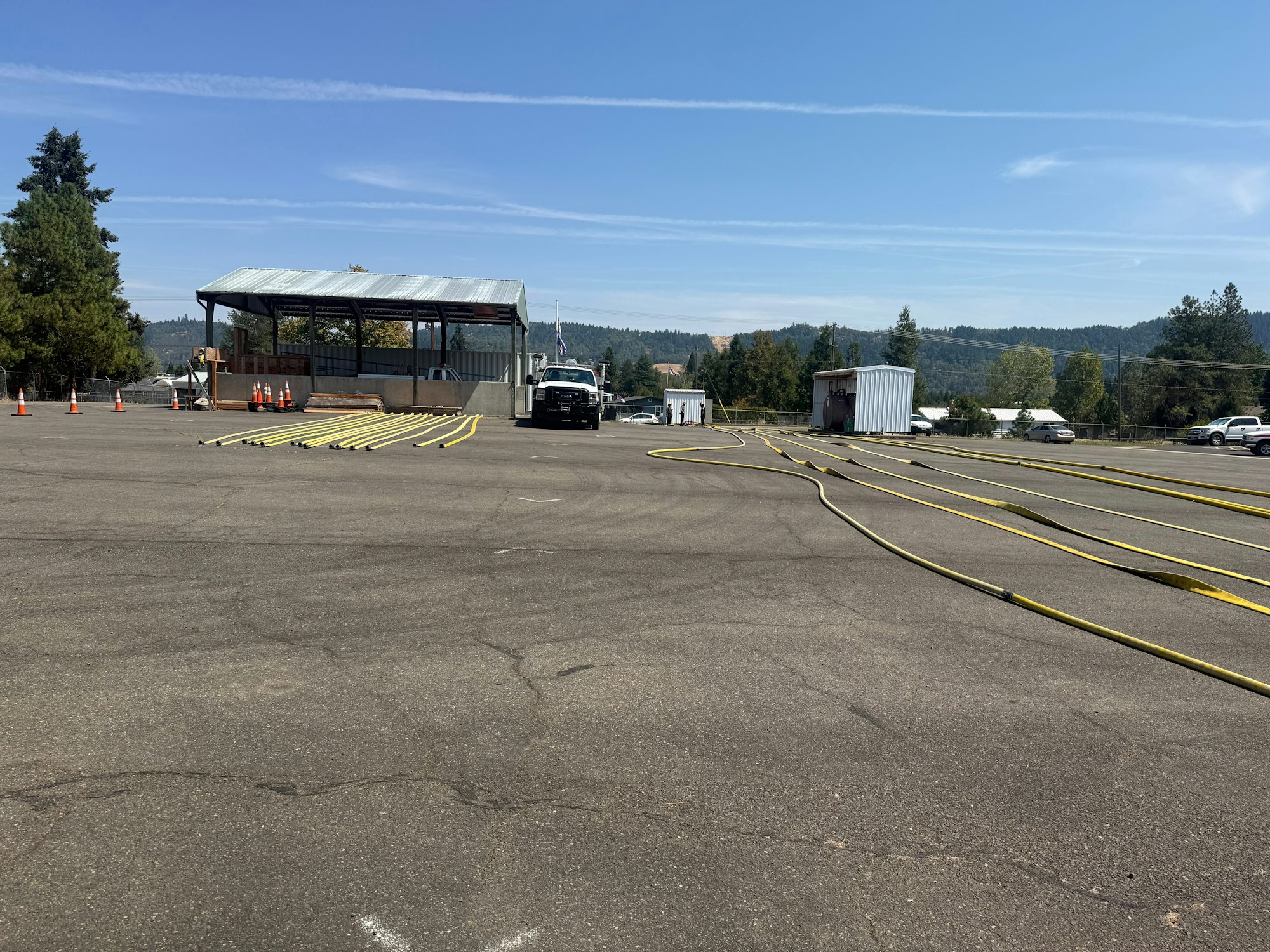 The image shows a spacious area with yellow hoses laid out, a structure, and parked vehicles under a clear blue sky.
