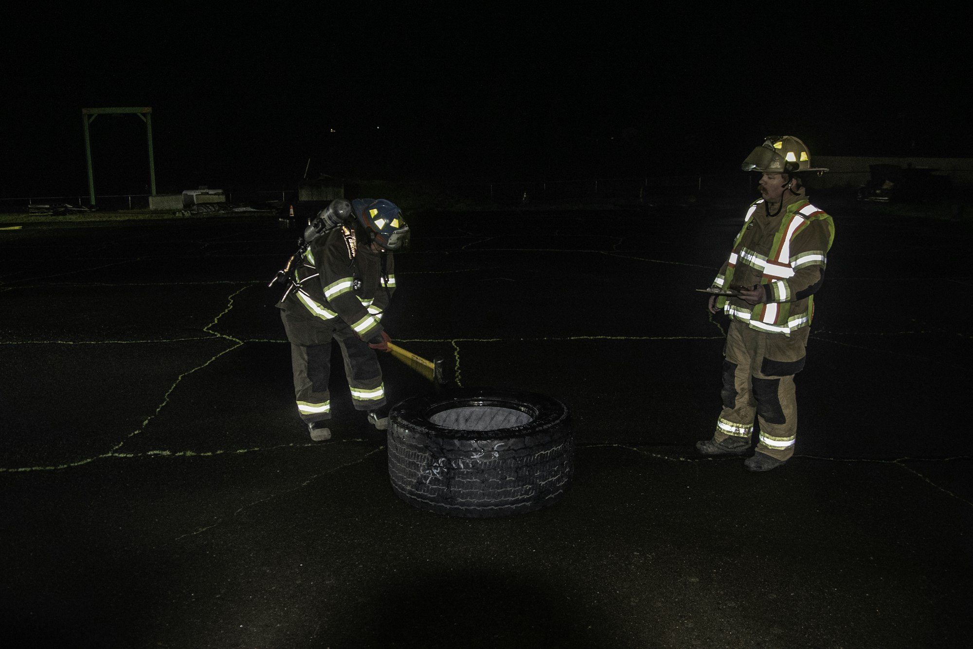 Two firefighters are working at night, one using a tool on a tire while the other observes and takes notes.