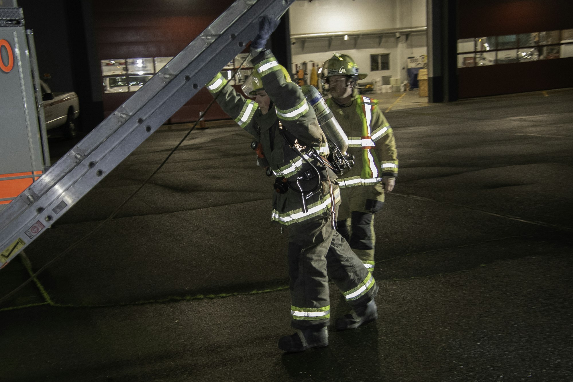 Two firefighters in gear are maneuvering a ladder in an outdoor setting, likely at a fire station or training area during the night.