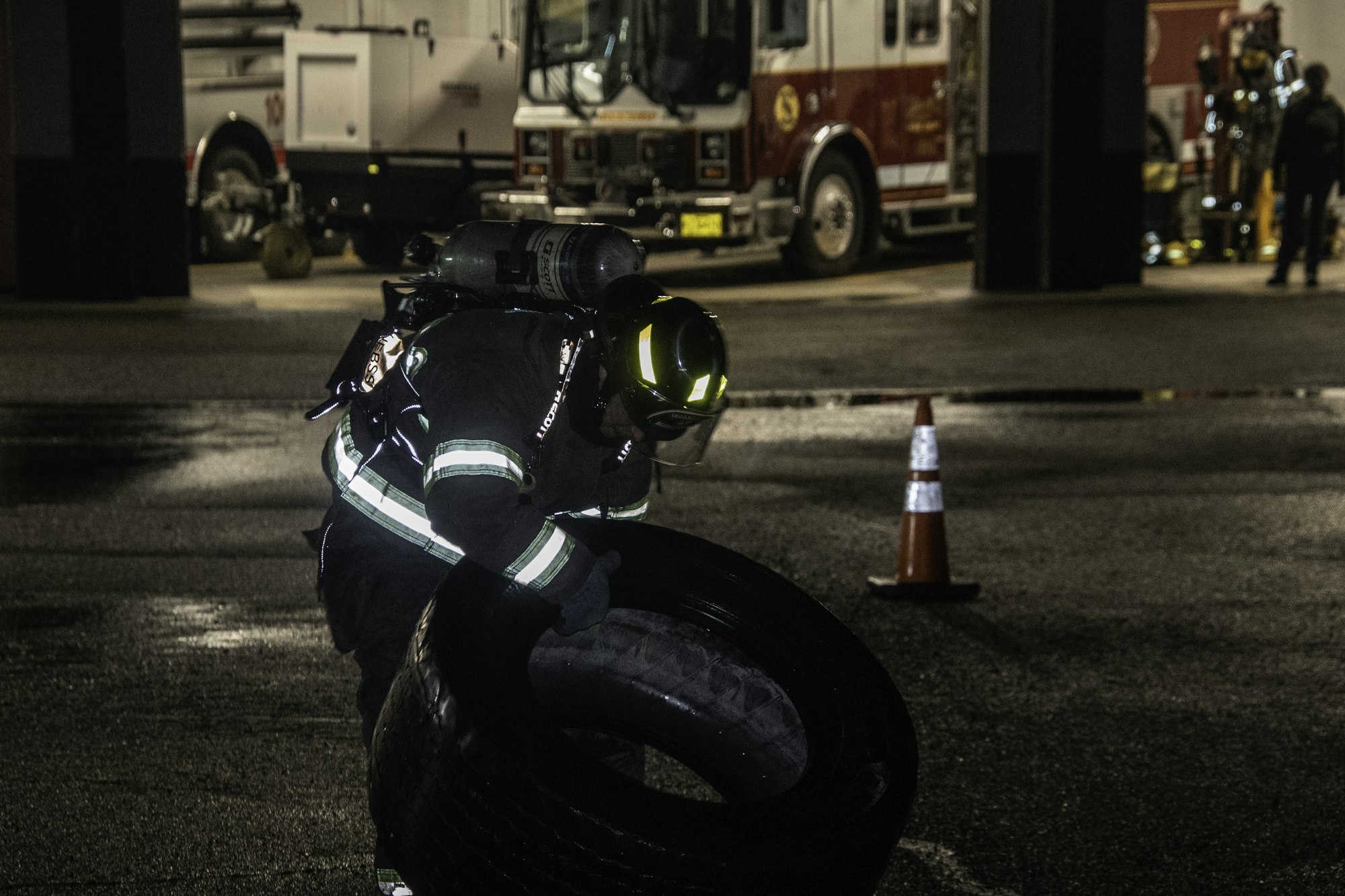 A firefighter in gear lifts a tire in a dimly lit garage, with fire trucks and equipment in the background.