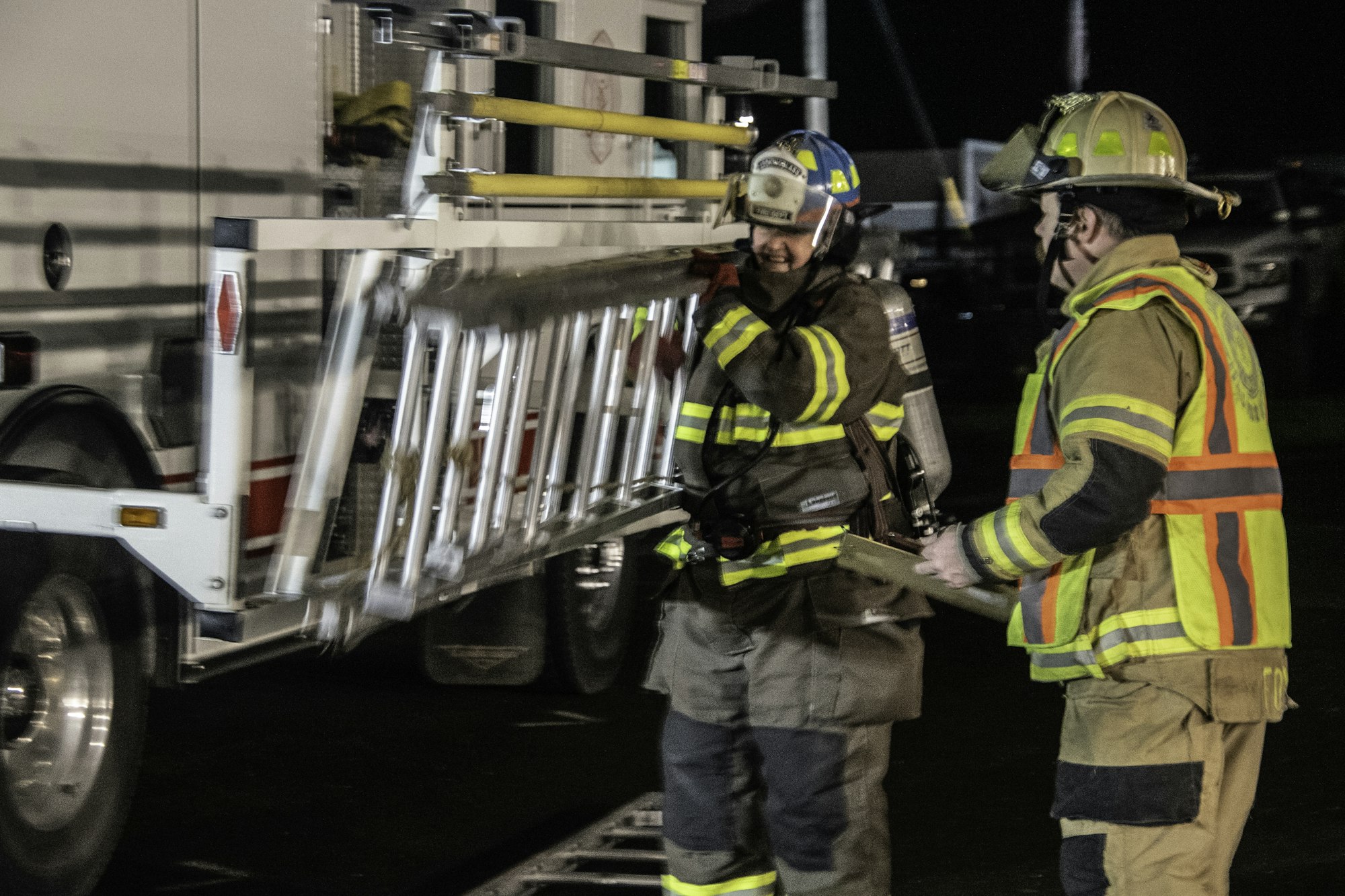Two firefighters are working together to lift a ladder from a fire truck at night. Safety gear is visible.
