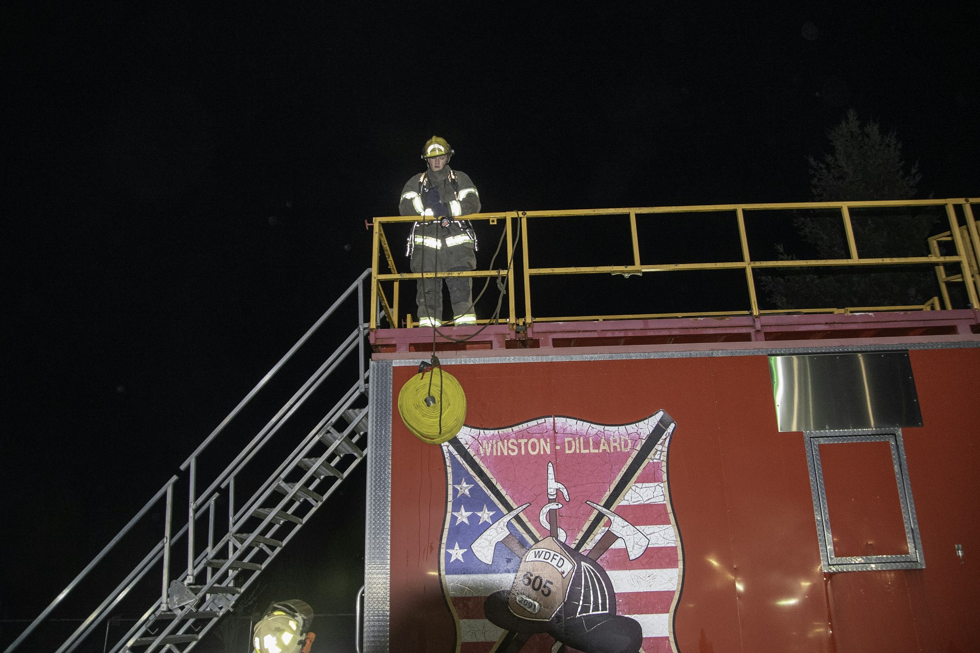 A firefighter stands on a roof platform at night, next to a bright red truck featuring a local fire department emblem.
