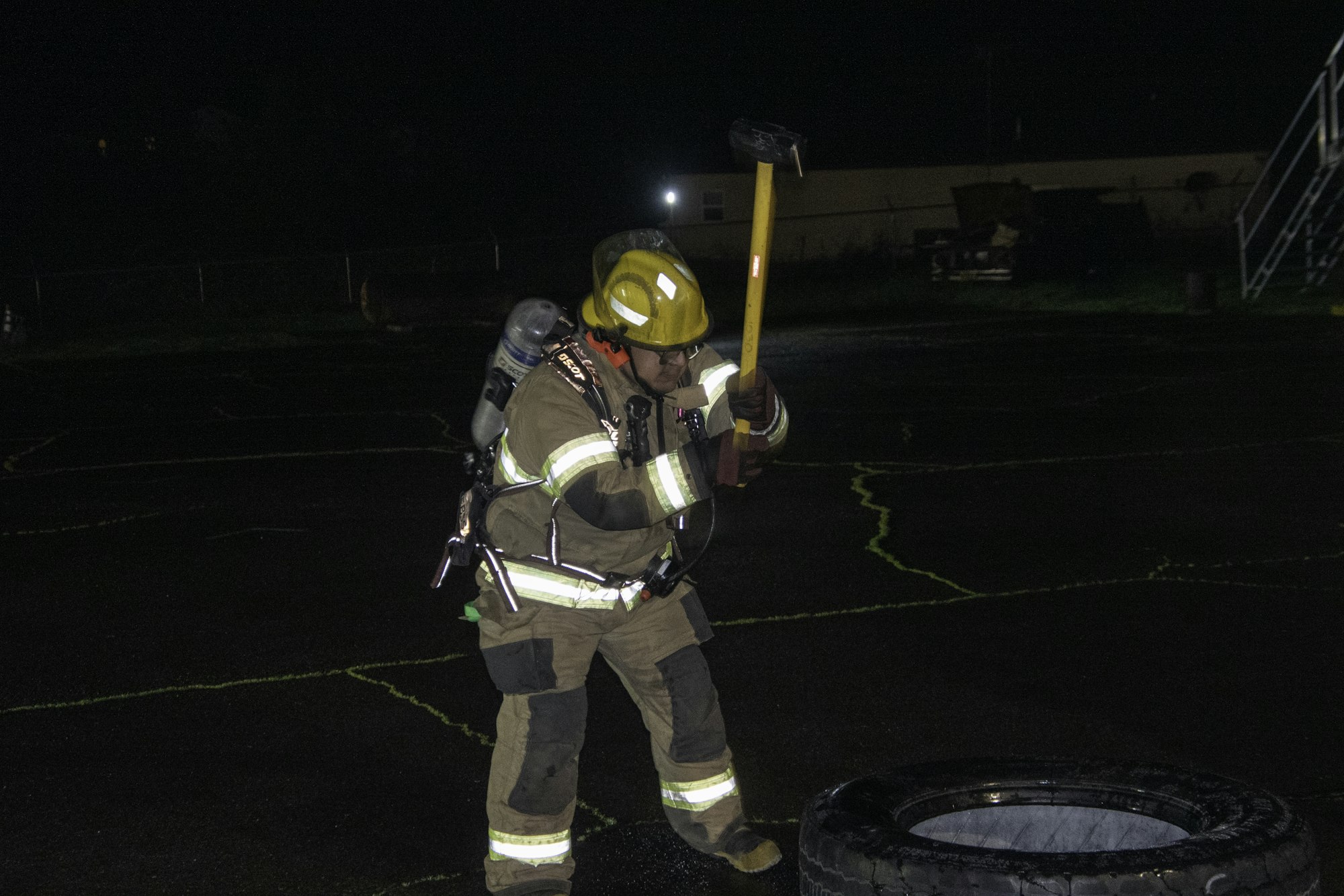 A firefighter in full gear is preparing to strike a tire with a sledgehammer at night.