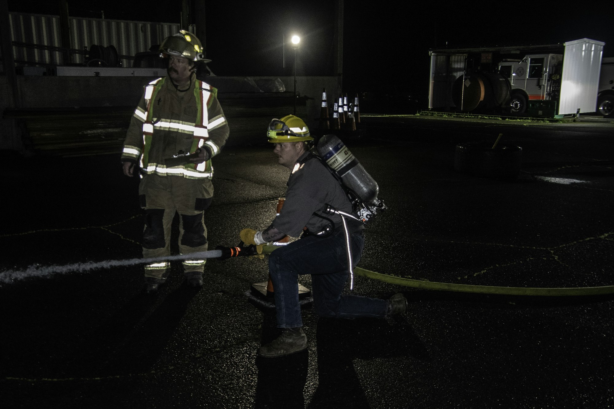 A firefighter training scene at night, with two individuals managing a fire hose and equipment in a dark environment.