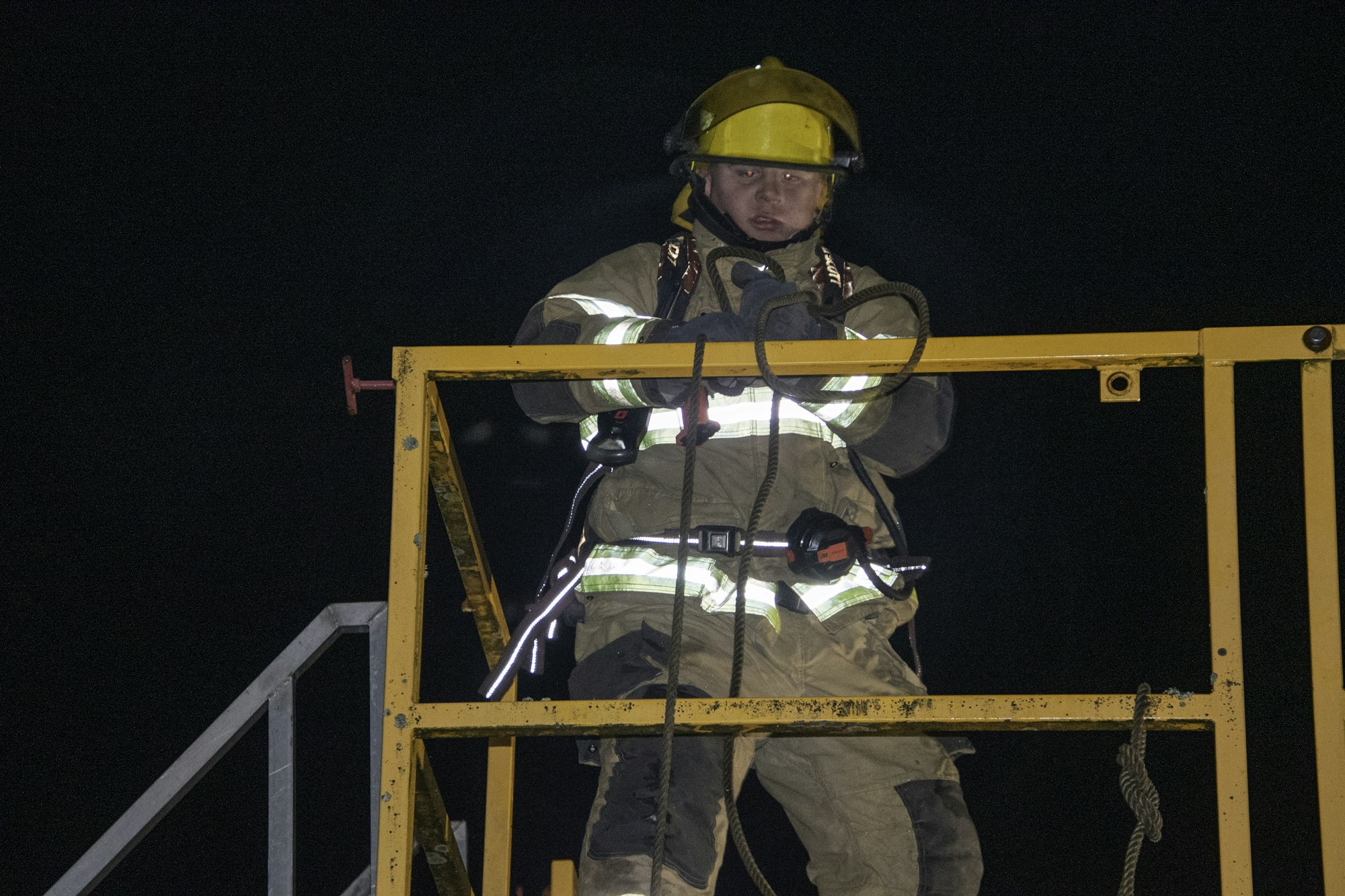 A firefighter in protective gear working on a platform at night, holding equipment and securing ropes.