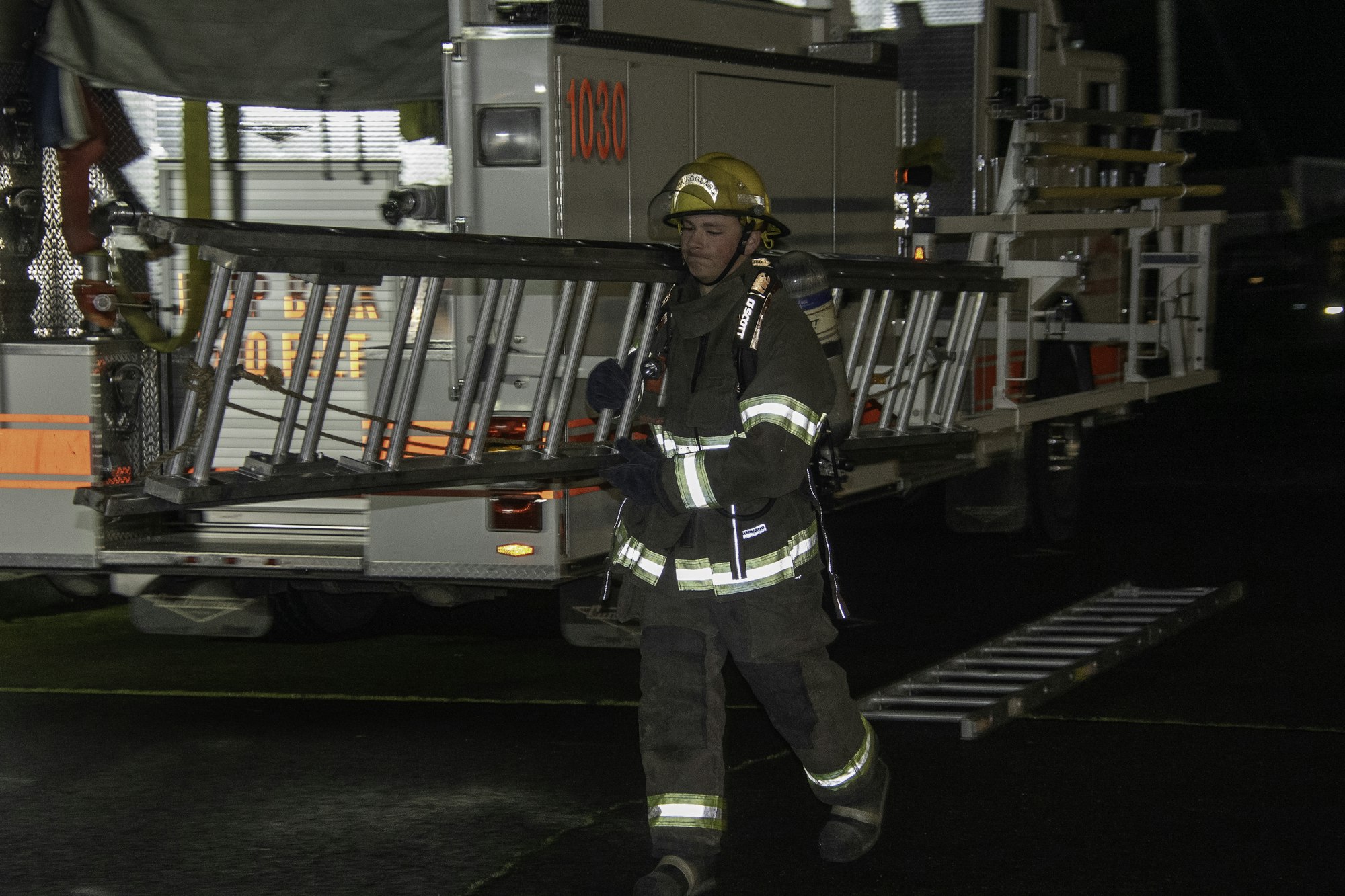 A firefighter carries a ladder away from a fire truck at night, preparing for emergency response.