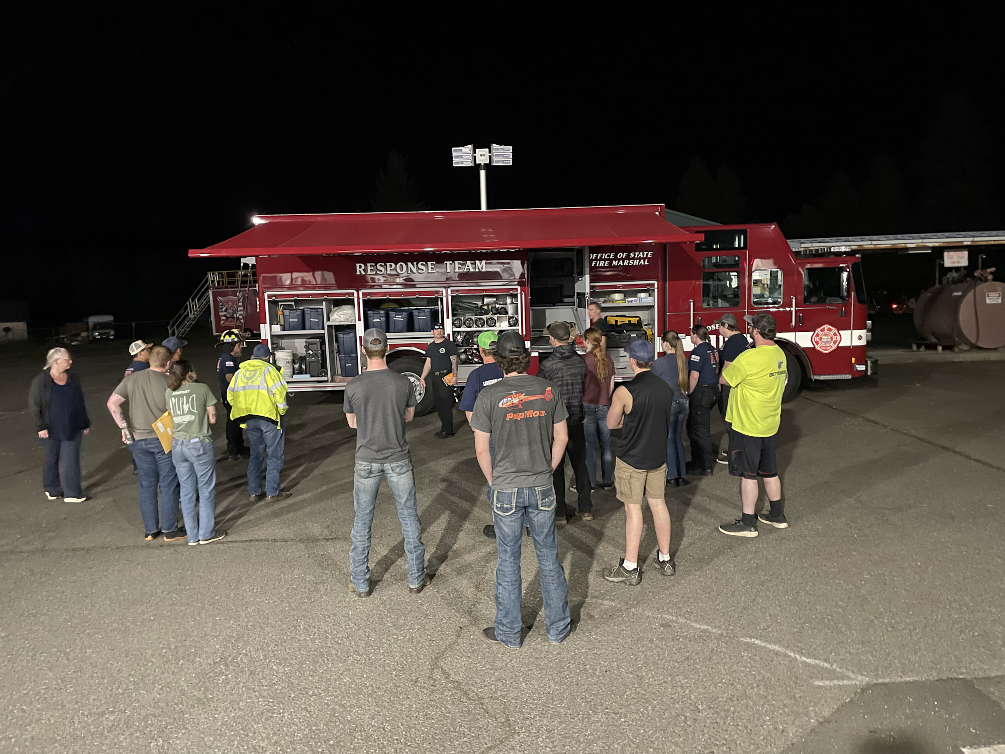 A group gathers around a red emergency response vehicle at night, possibly for a training or briefing session.