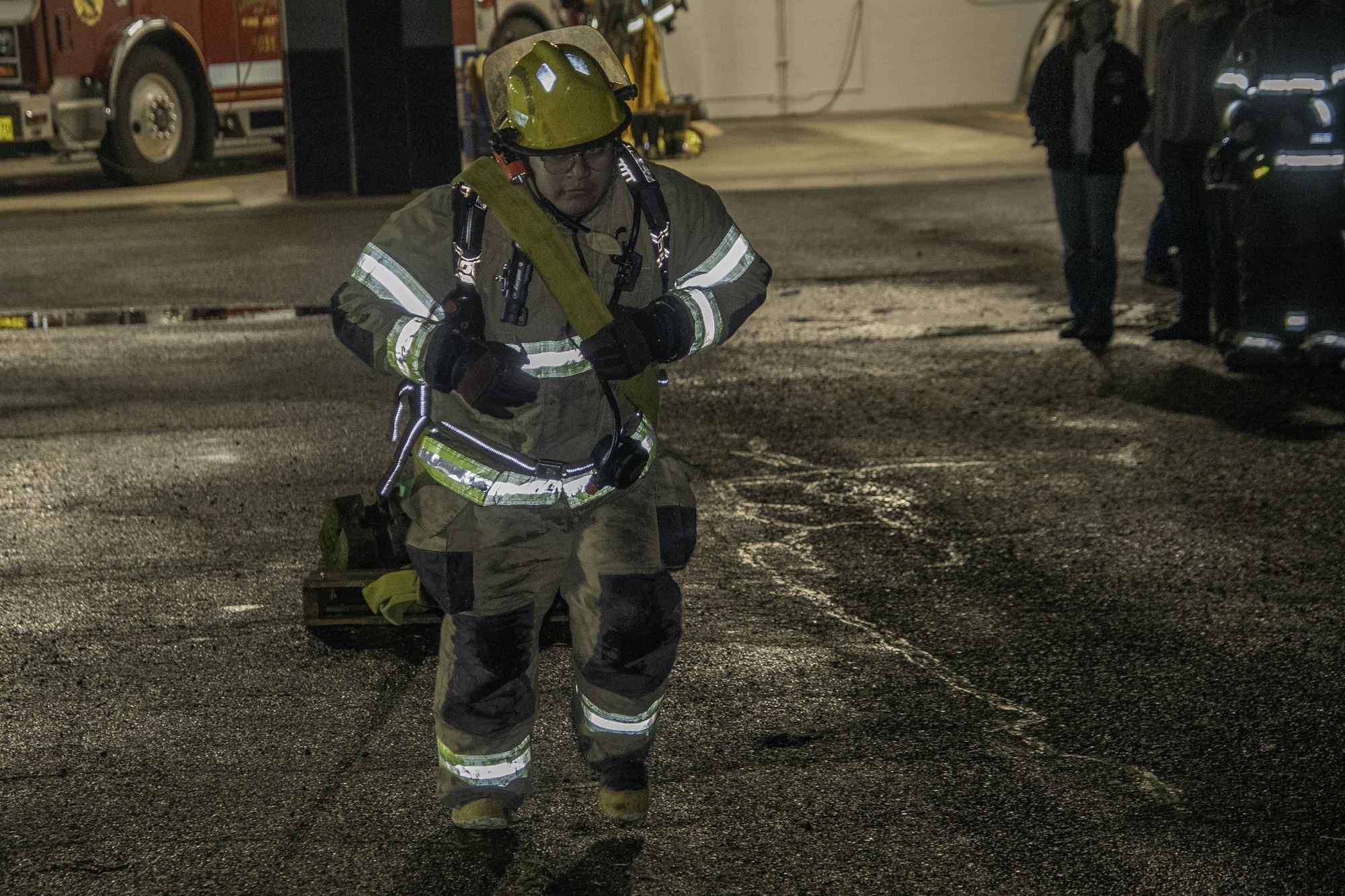 A firefighter in uniform, carrying equipment, walks on a wet surface near a fire truck and bystanders.