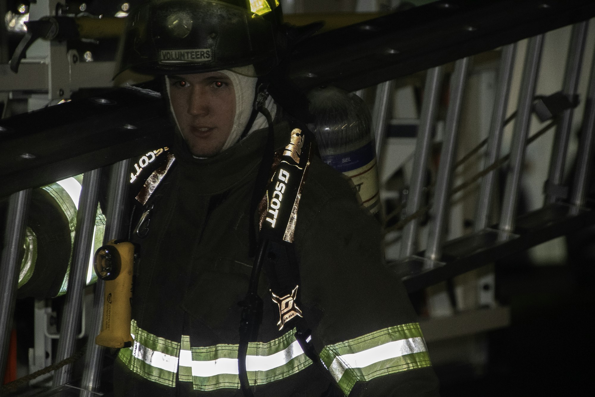 A firefighter in gear, standing next to a ladder, with safety equipment and a flashlight, ready for action.