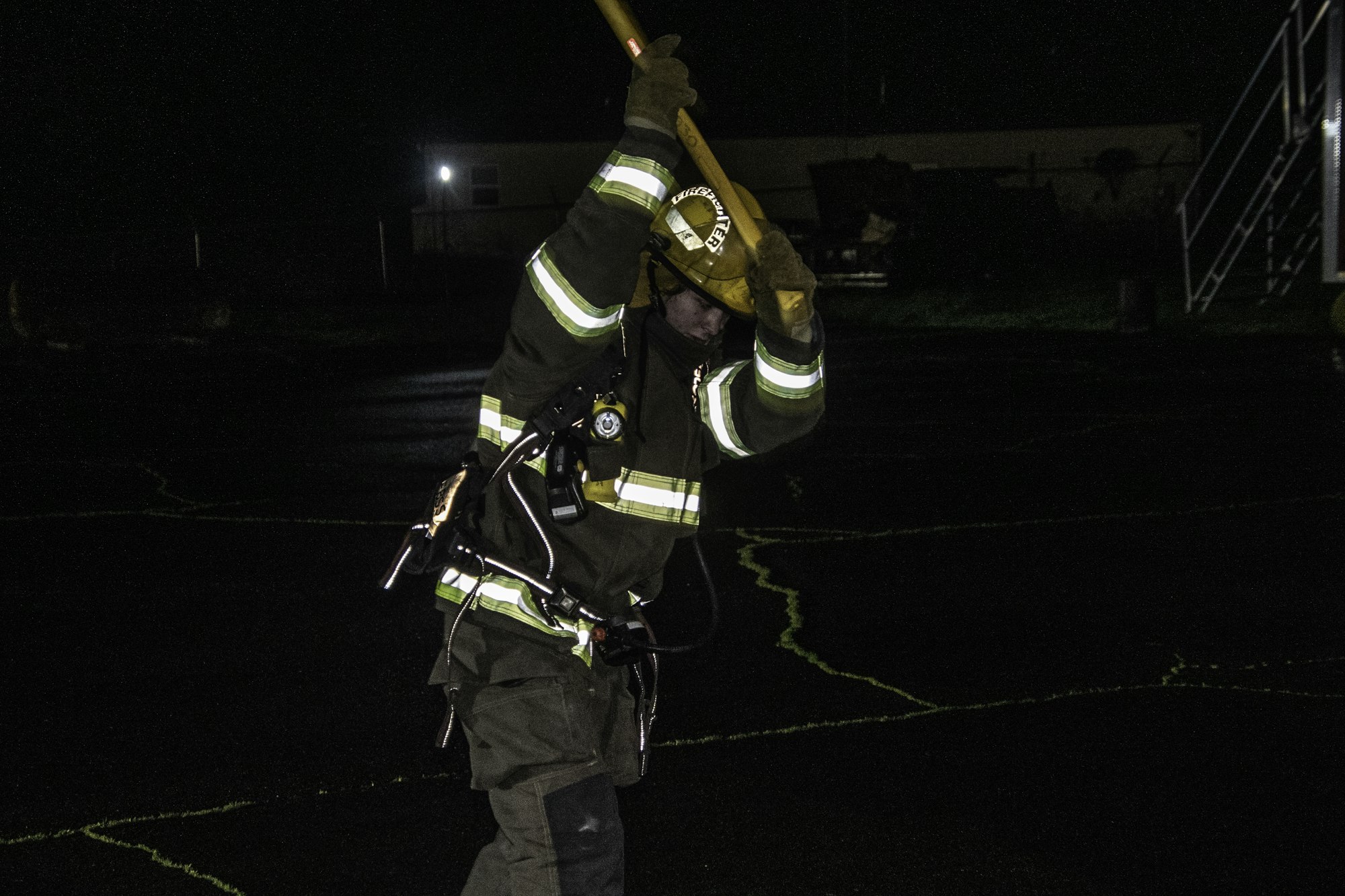 A firefighter in gear is swinging a tool, possibly during a training exercise, in a dark outdoor setting.