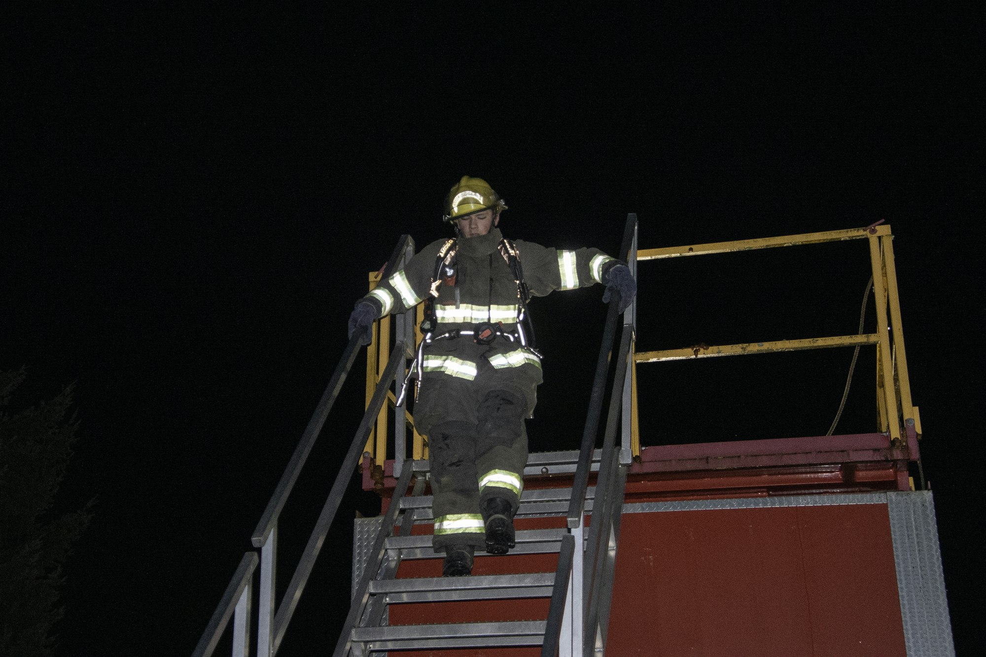 A firefighter in gear descends a metallic staircase at night, illuminated by equipment.