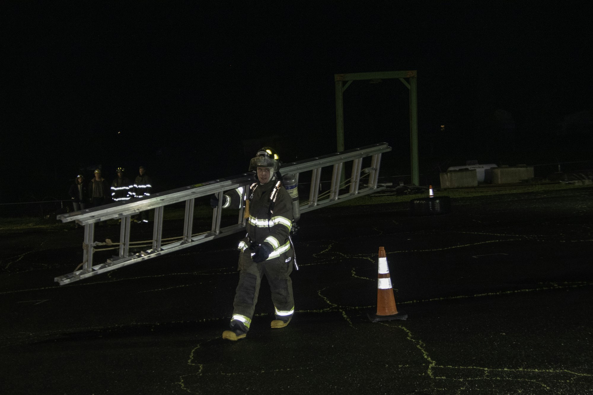 A firefighter is carrying a ladder at night, with other emergency personnel visible in the background.