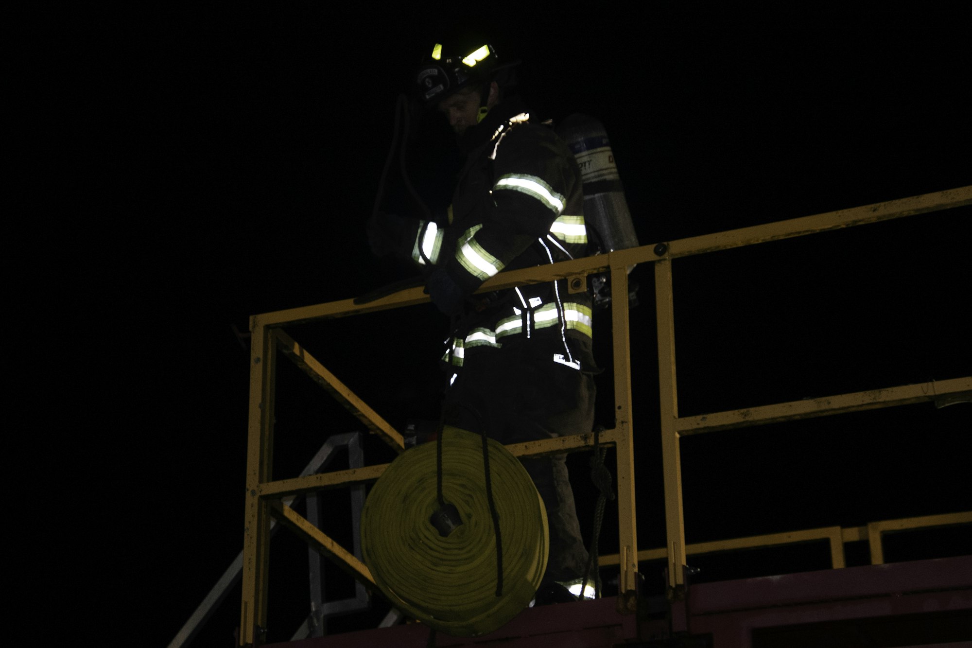 A firefighter is atop a platform at night, holding a harness and standing next to a coiled hose.