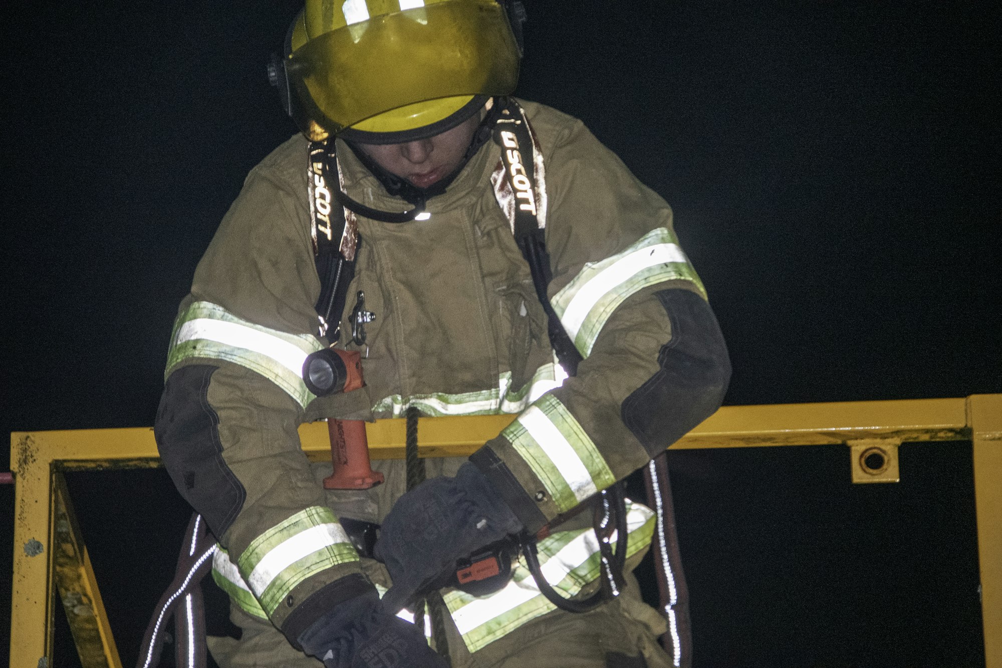A firefighter in gear, wearing a helmet, works on a harness or equipment at night, focused on the task at hand.