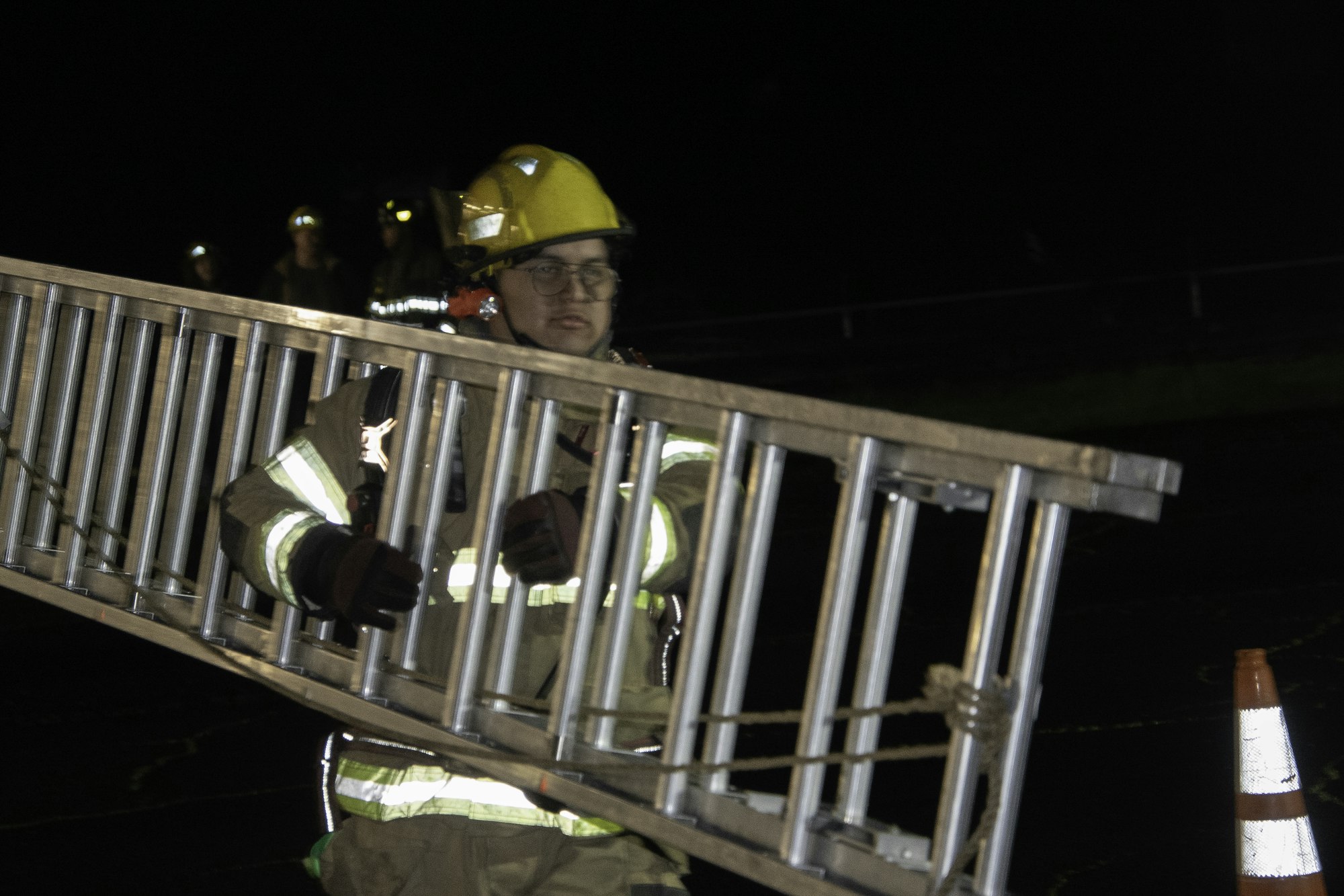 A firefighter in gear carries a ladder at night, with other firefighters visible in the background.