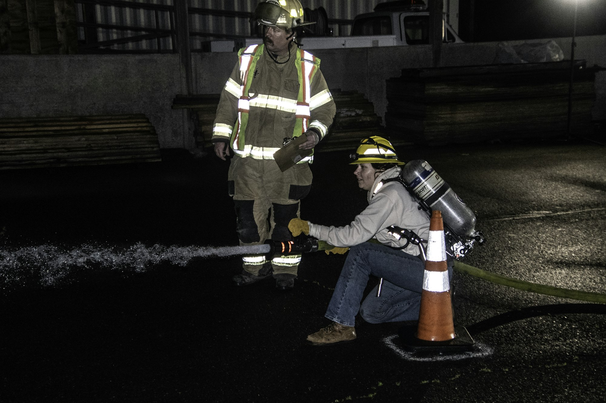 A firefighter trains at night, using a hose to spray water while another supervises, ensuring safety and technique.