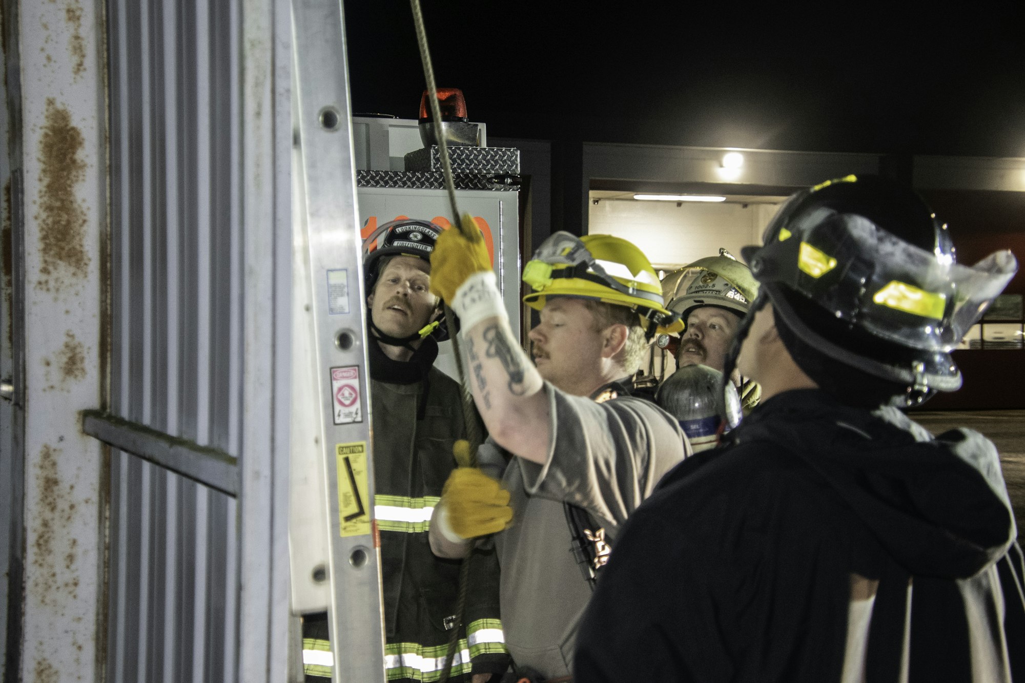 Firefighters are gathered, focused on a task involving a ladder and equipment at night, showcasing teamwork and safety preparation.