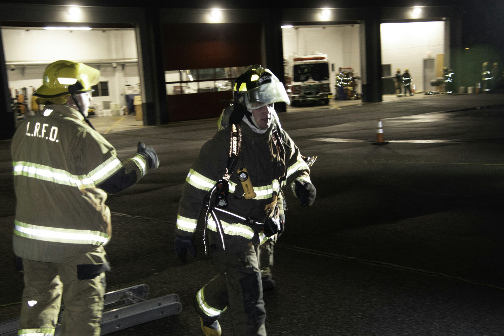 Two firefighters in gear are maneuvering outside a fire station at night, with emergency vehicles visible in the background.