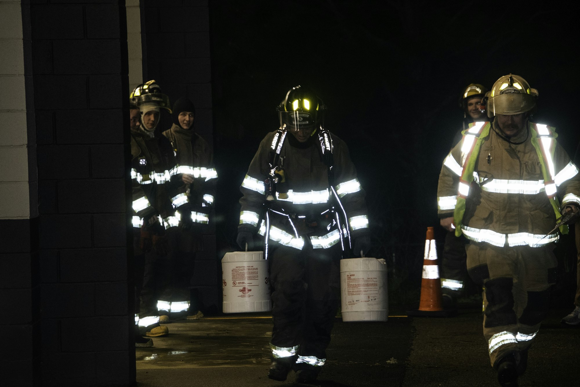 Firefighters in protective gear carrying buckets, working at night, with others standing nearby. Safety cones visible.