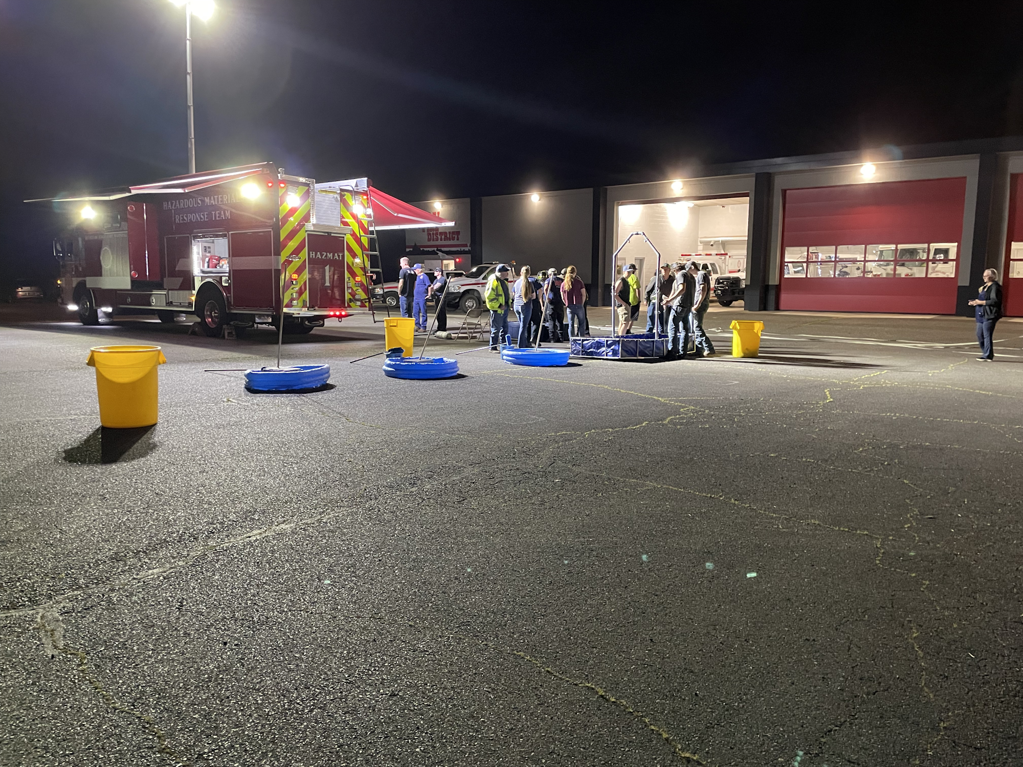 A night scene showing a hazardous materials response team with a fire truck, gathering around equipment in a parking lot.