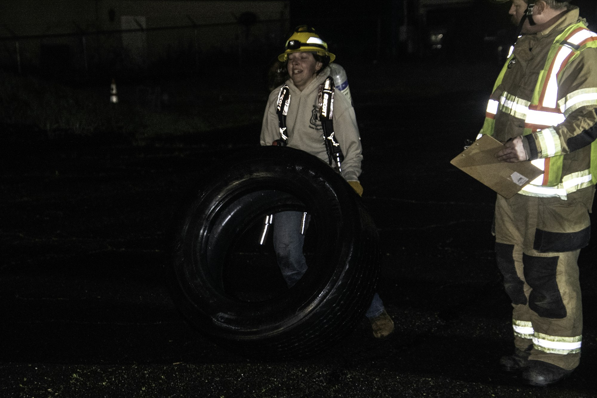 A person wearing a helmet carries a large tire while another person in safety gear holds a clipboard, in a dark setting.