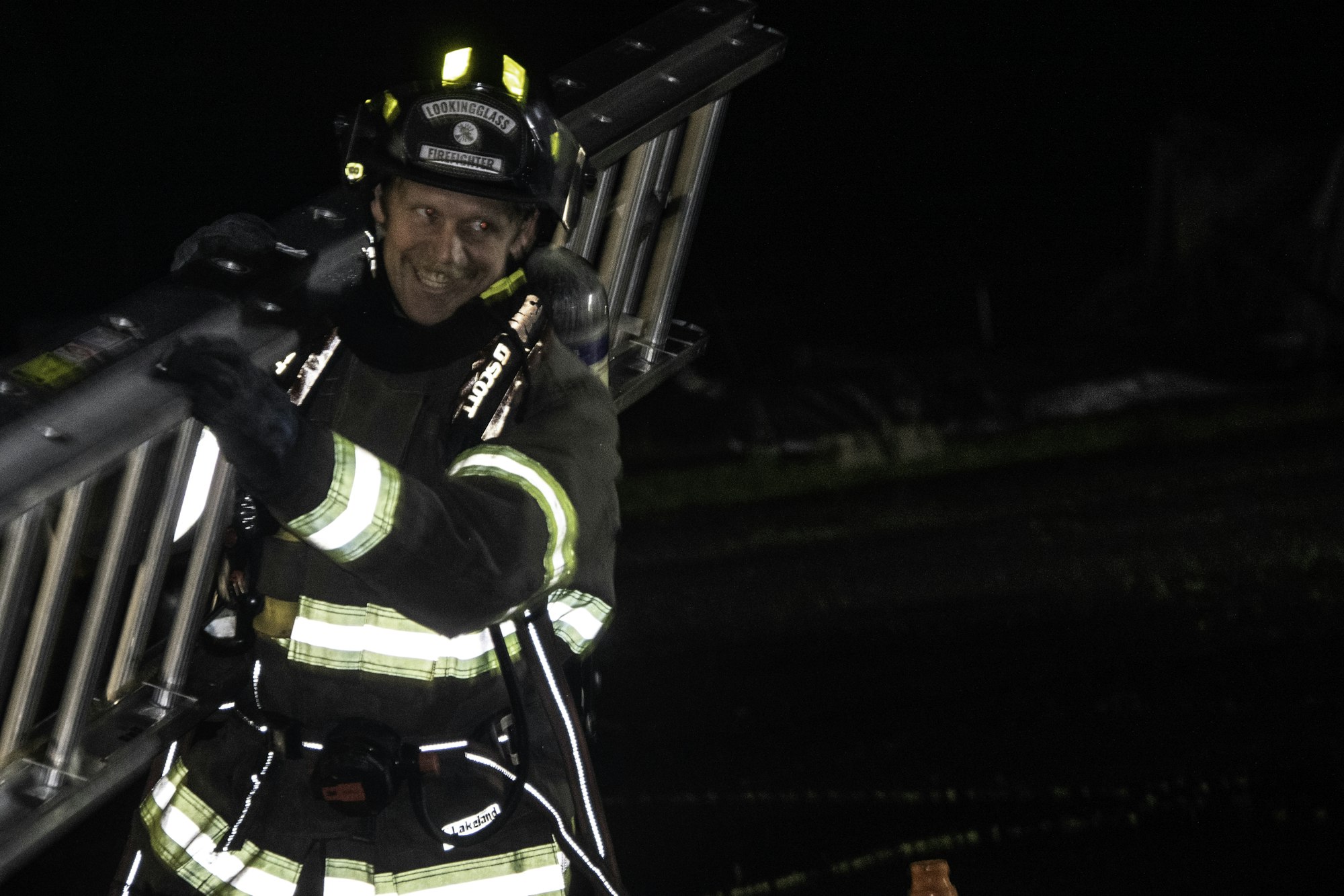 A firefighter in gear smiling while carrying a ladder at night, demonstrating readiness and teamwork in emergency response.