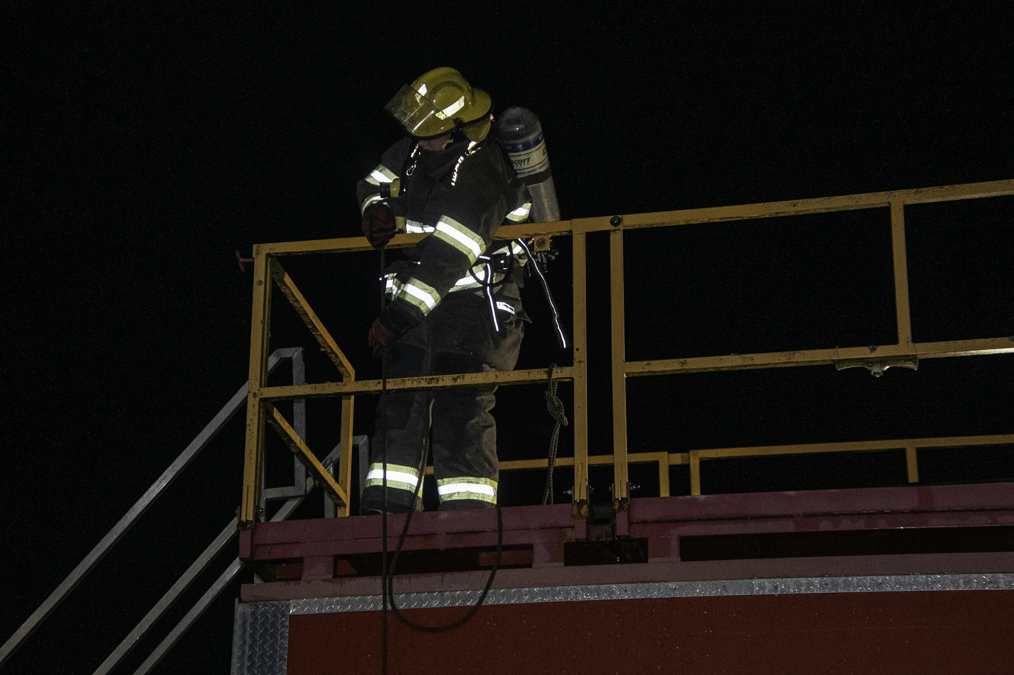 A firefighter in gear stands on a raised platform at night, preparing equipment for a task or training. Safety measures visible.