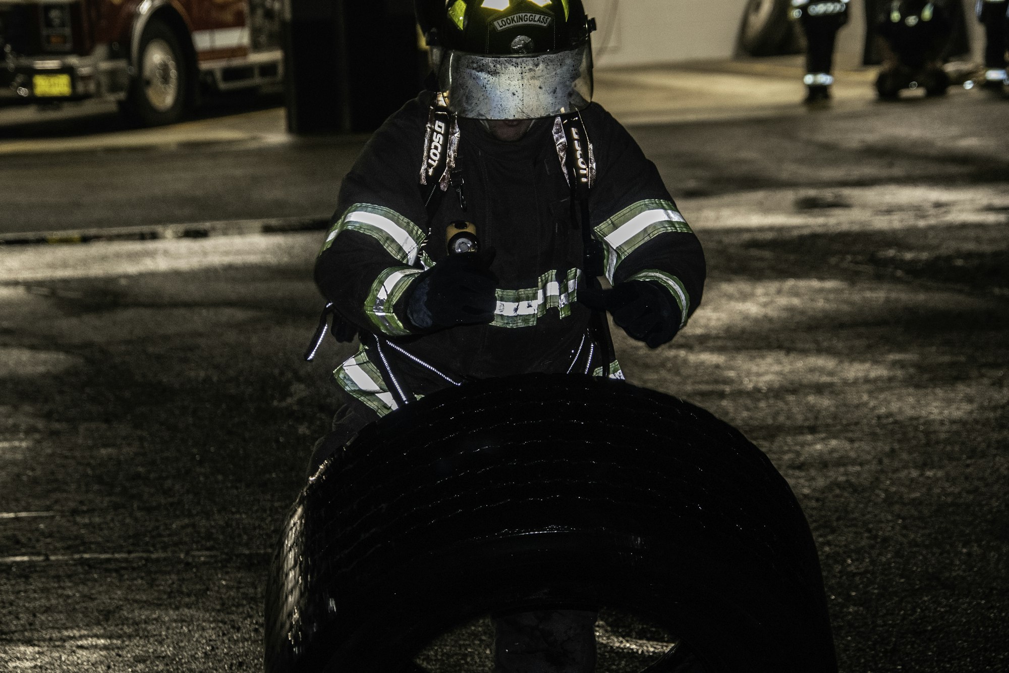 A firefighter in gear is handling a large tire in a dimly lit environment, possibly during training or a drill.